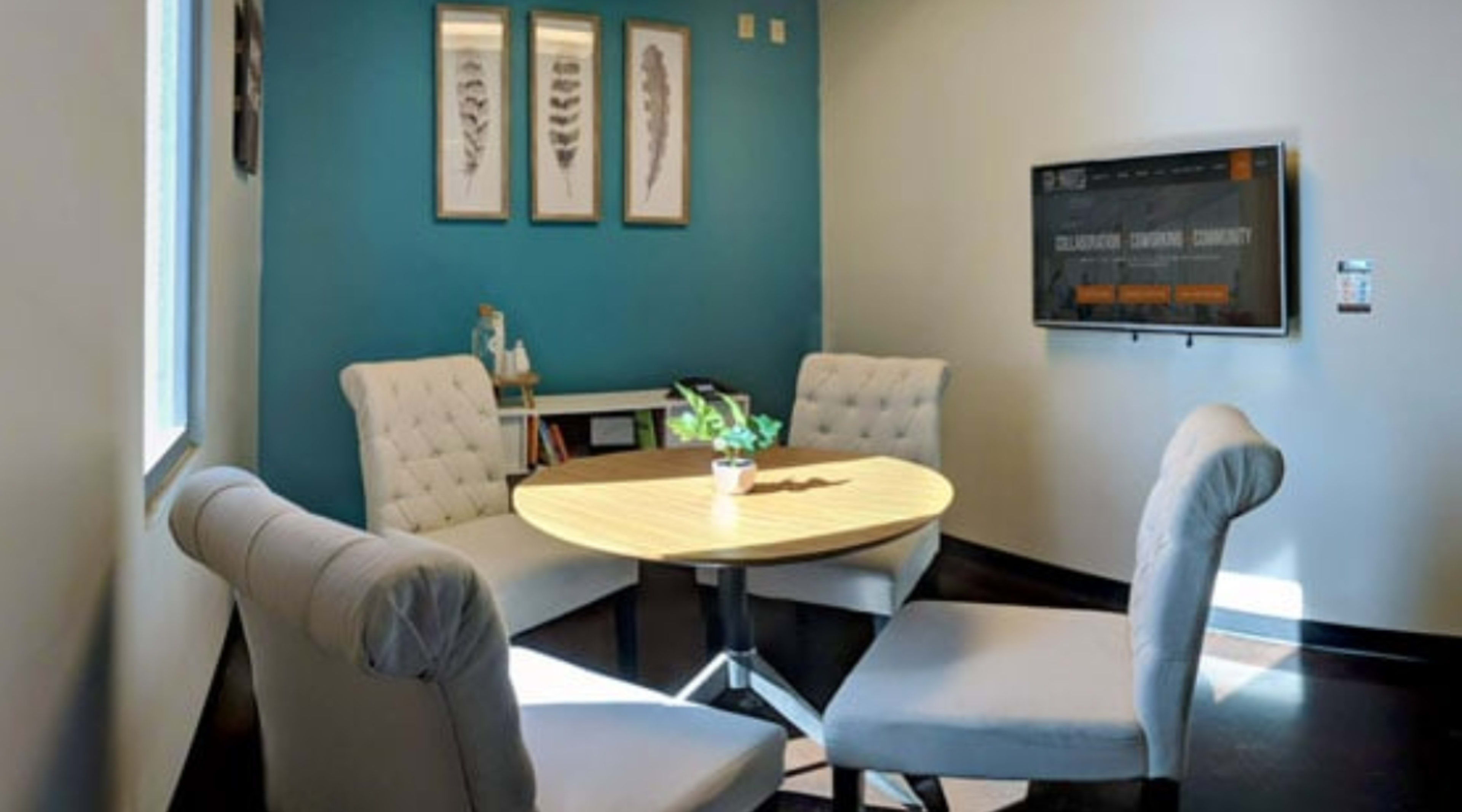 A small meeting room features a round table surrounded by four upholstered chairs, with blue walls and framed botanical prints.