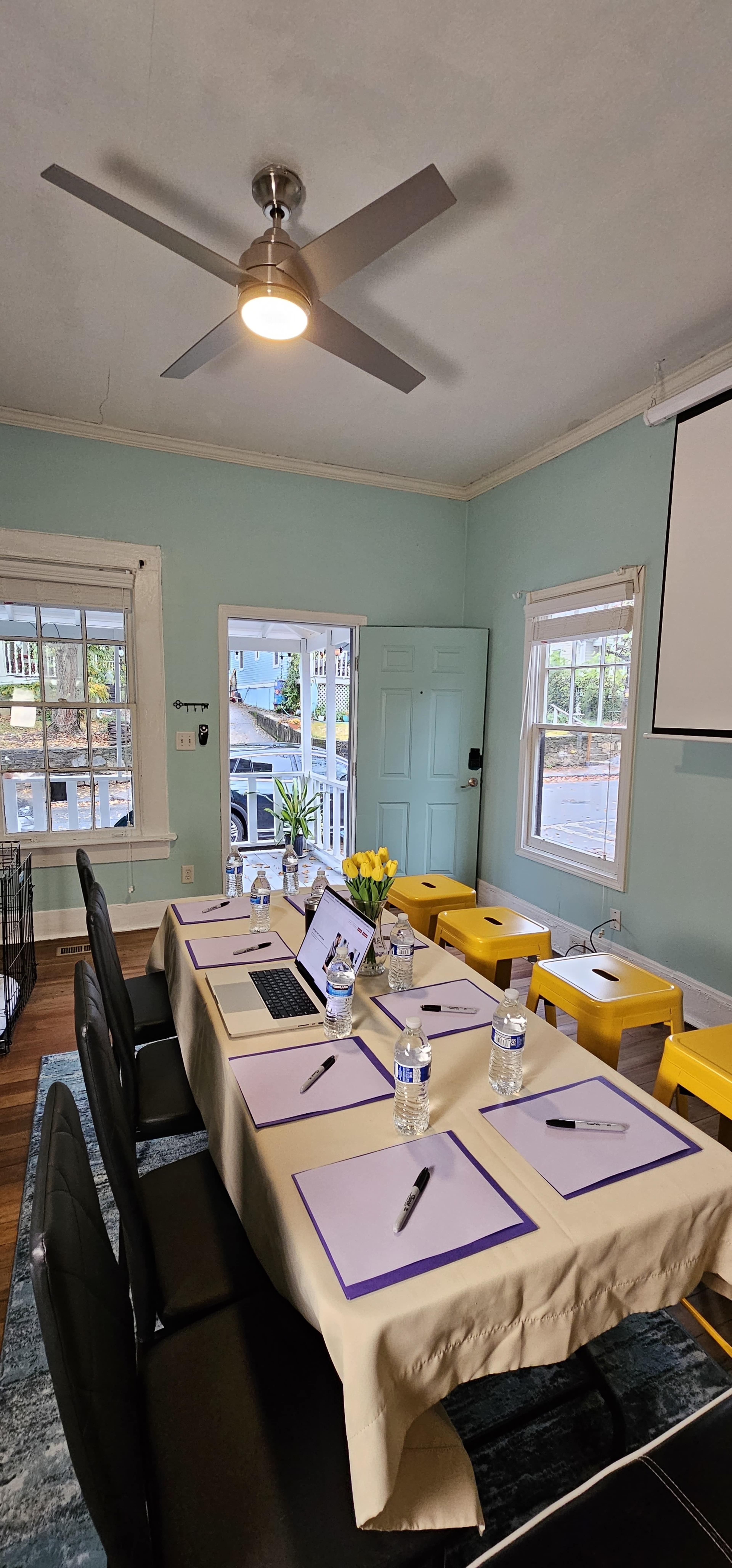 A meeting room is set up with a long table covered in a beige cloth, equipped with laptops, water bottles, and notepads, while large windows let in natural light.