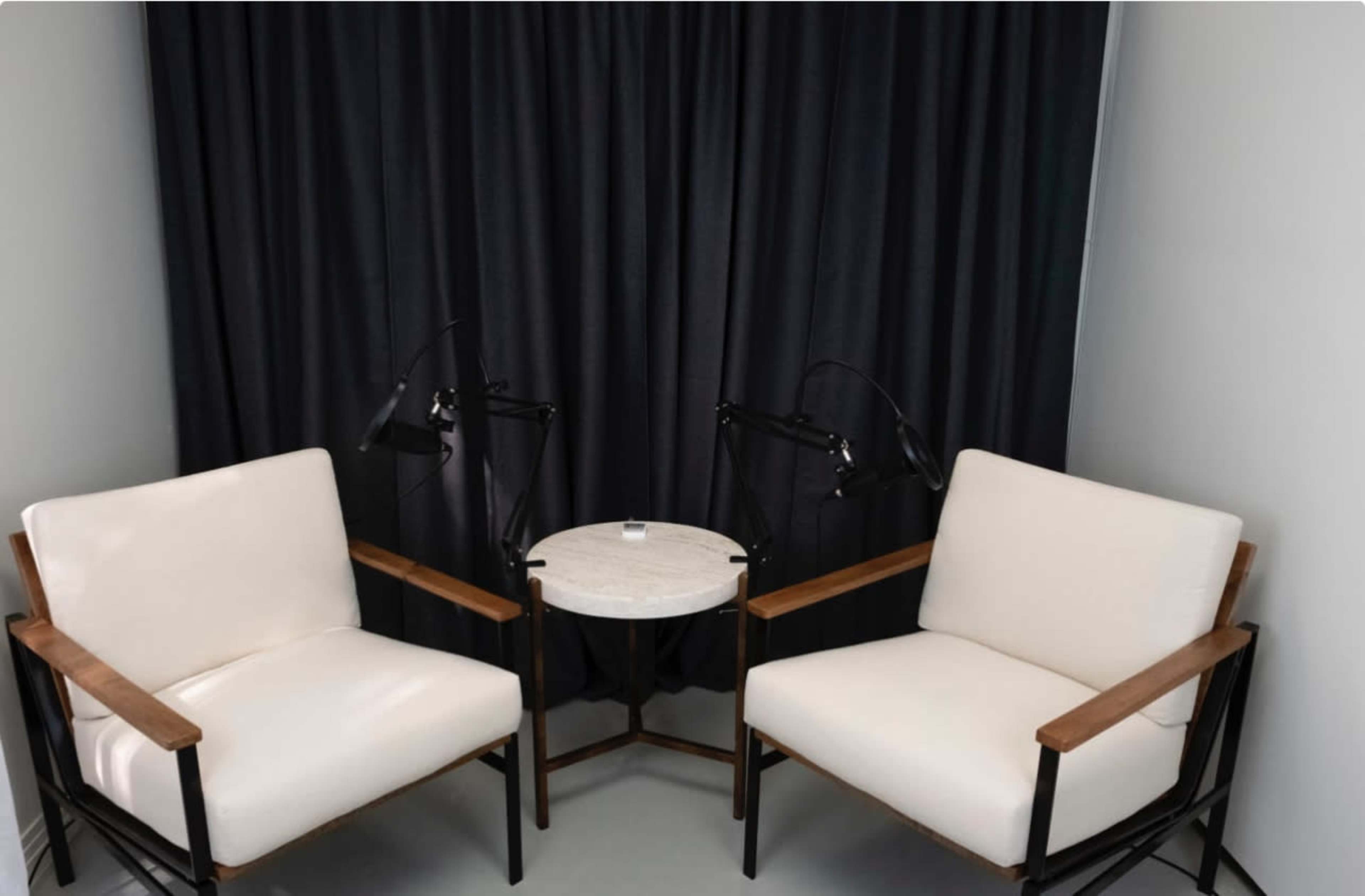 Two white armchairs with wooden accents face a small round table, positioned in front of a black curtain.