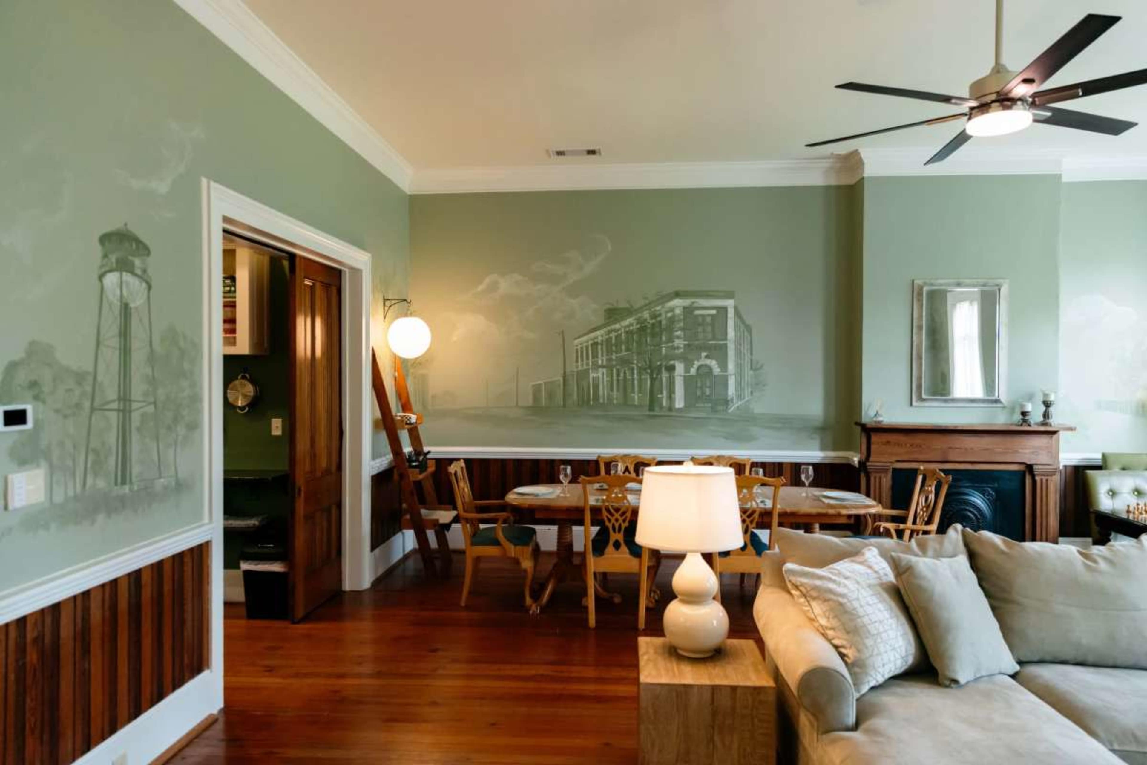 The image shows a stylish interior living space featuring a mural on the wall, a dining area with wooden furniture, and a cozy seating area with a light-colored sofa and a table lamp.
