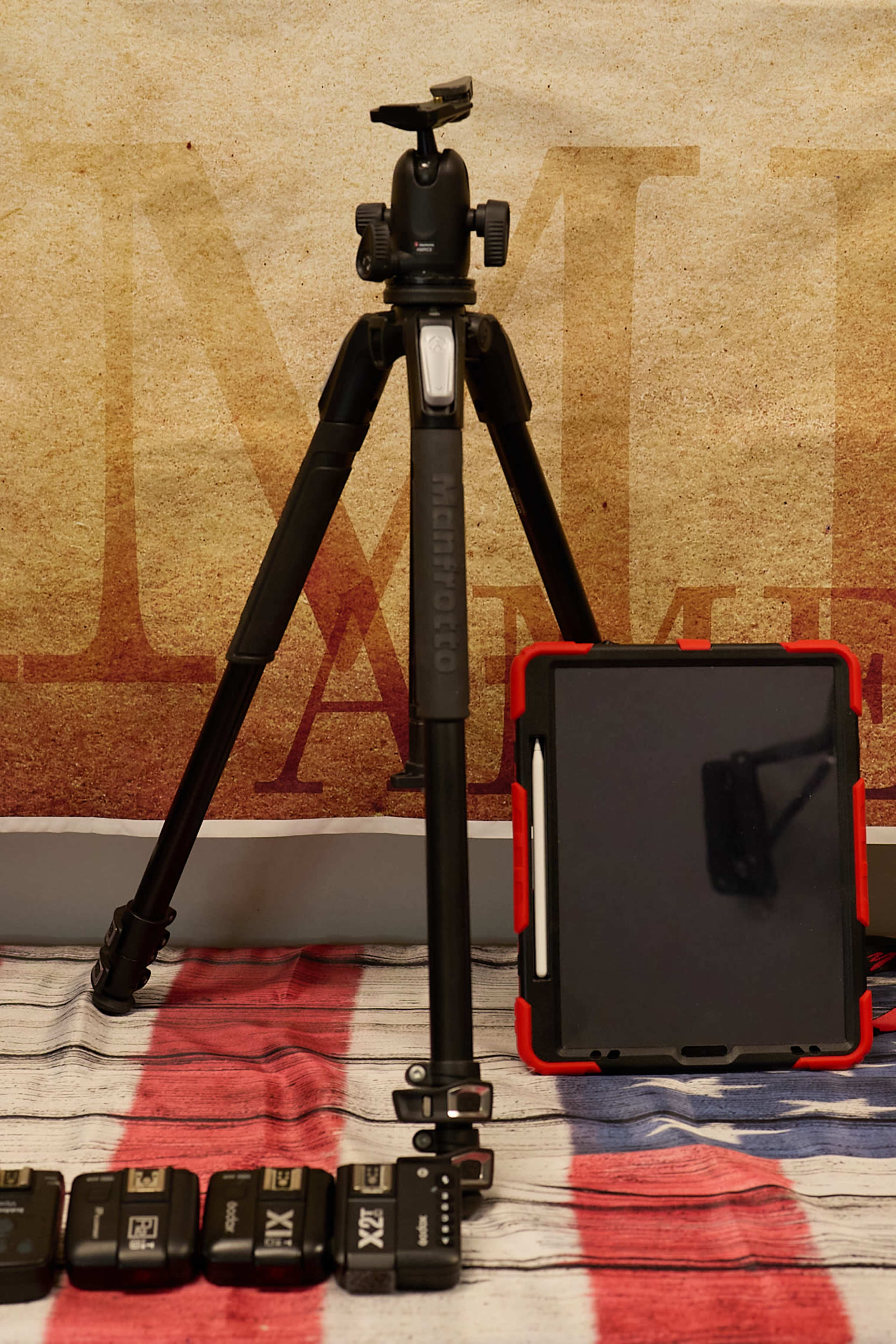 A black tripod stands next to a red-cased tablet and several camera batteries lined up on a table.
