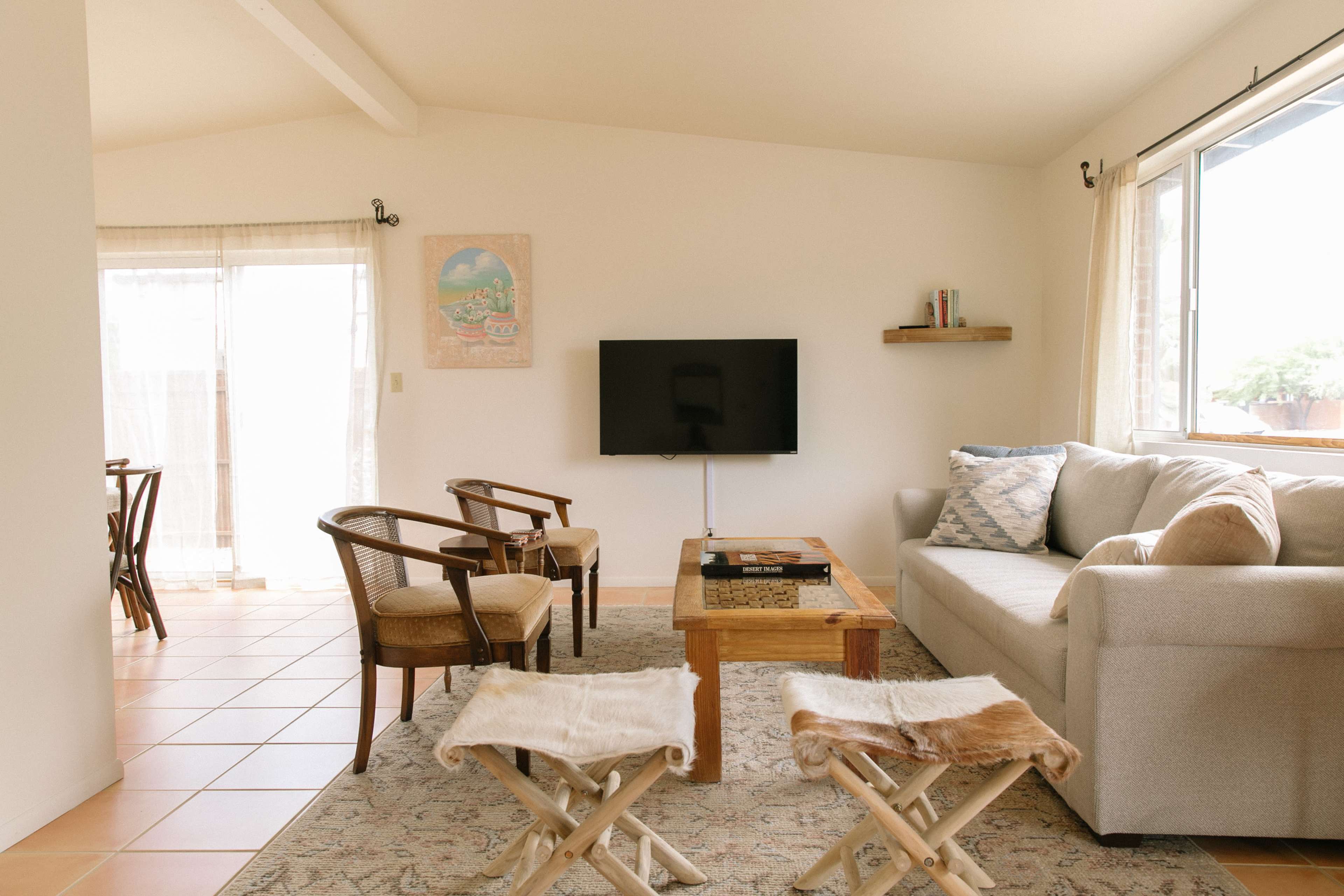 A bright living room featuring a gray sofa, a wooden coffee table, two chairs, and a television mounted on the wall, with a view of a dining area and large windows.