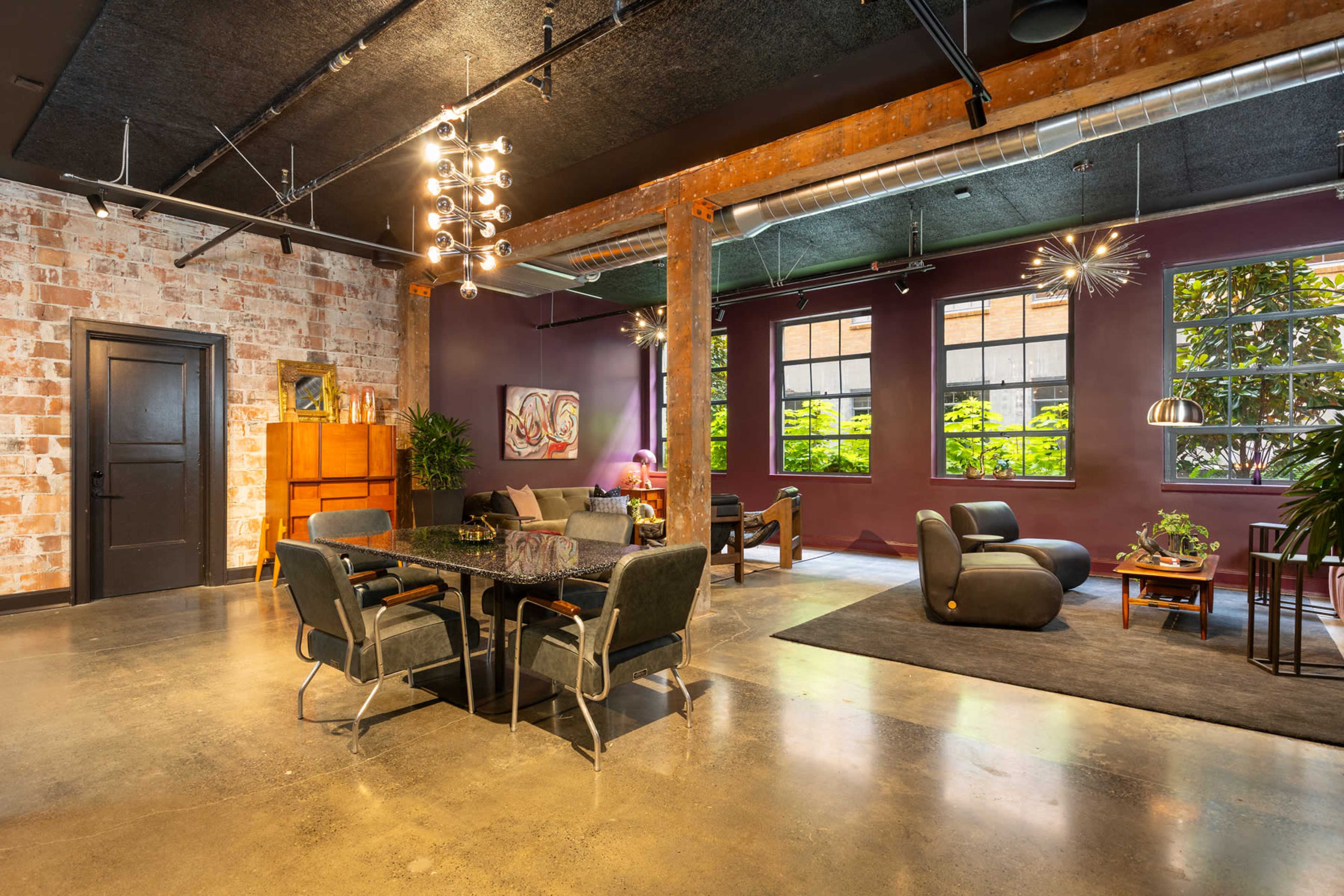 A spacious, modern loft interior with exposed brick walls, large windows, and a mix of seating areas, including a dining table and a lounge space.
