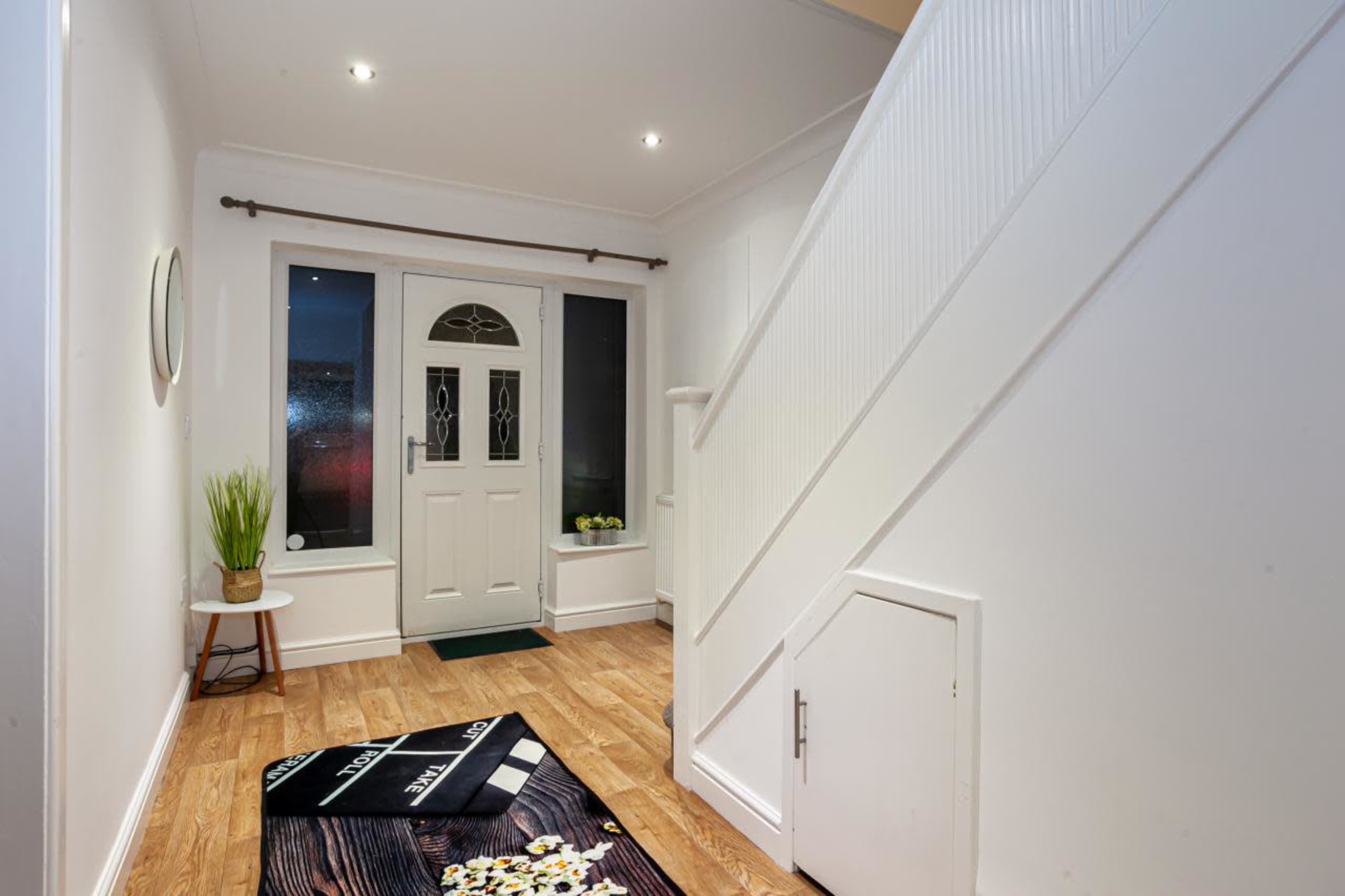 A hallway with a white door, staircase, and a wooden floor, featuring a decorative area rug and a small plant on a table.