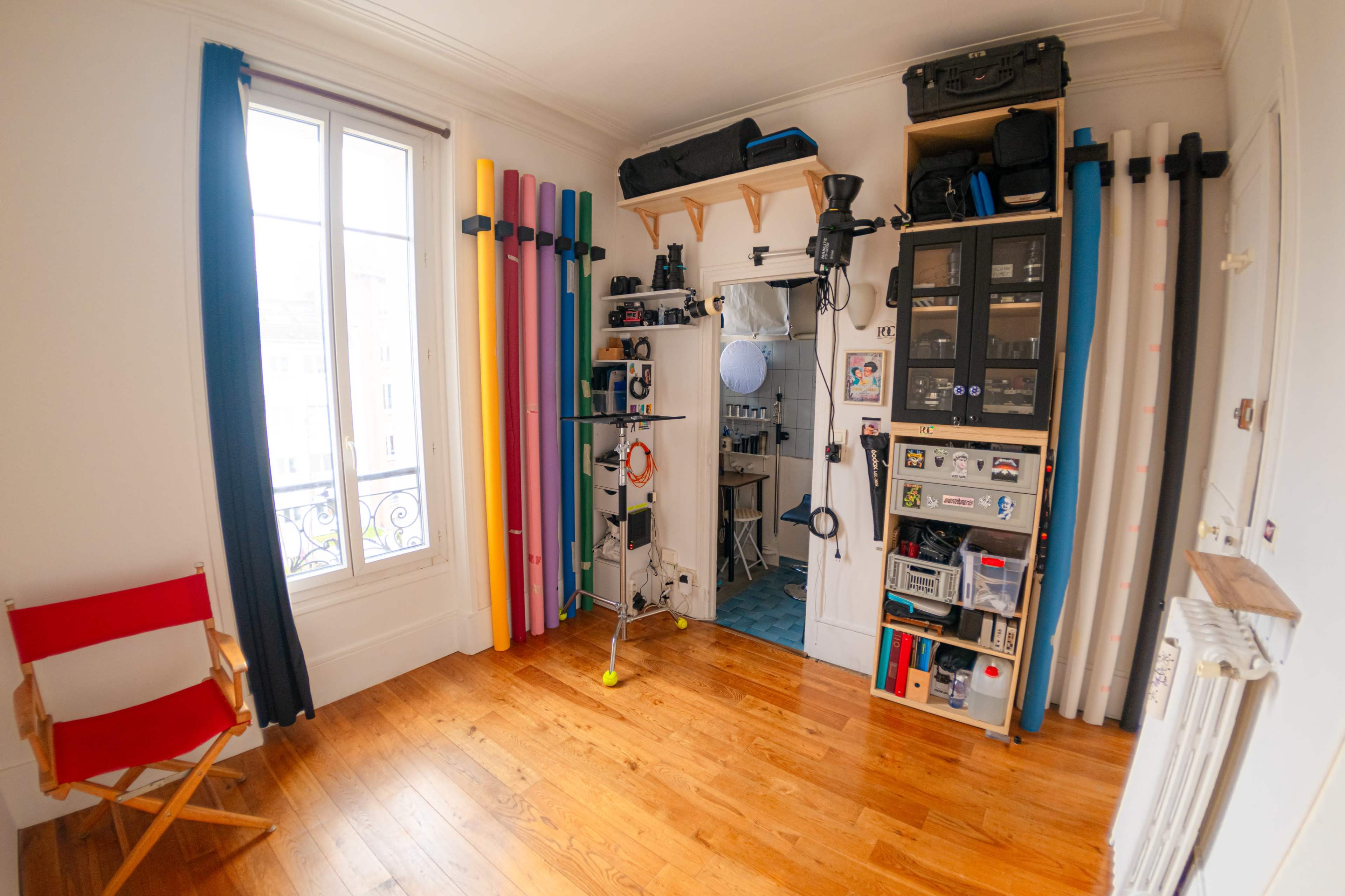 A room features a mix of colorful photography backdrops, storage shelves, and a chair, with large windows allowing natural light.