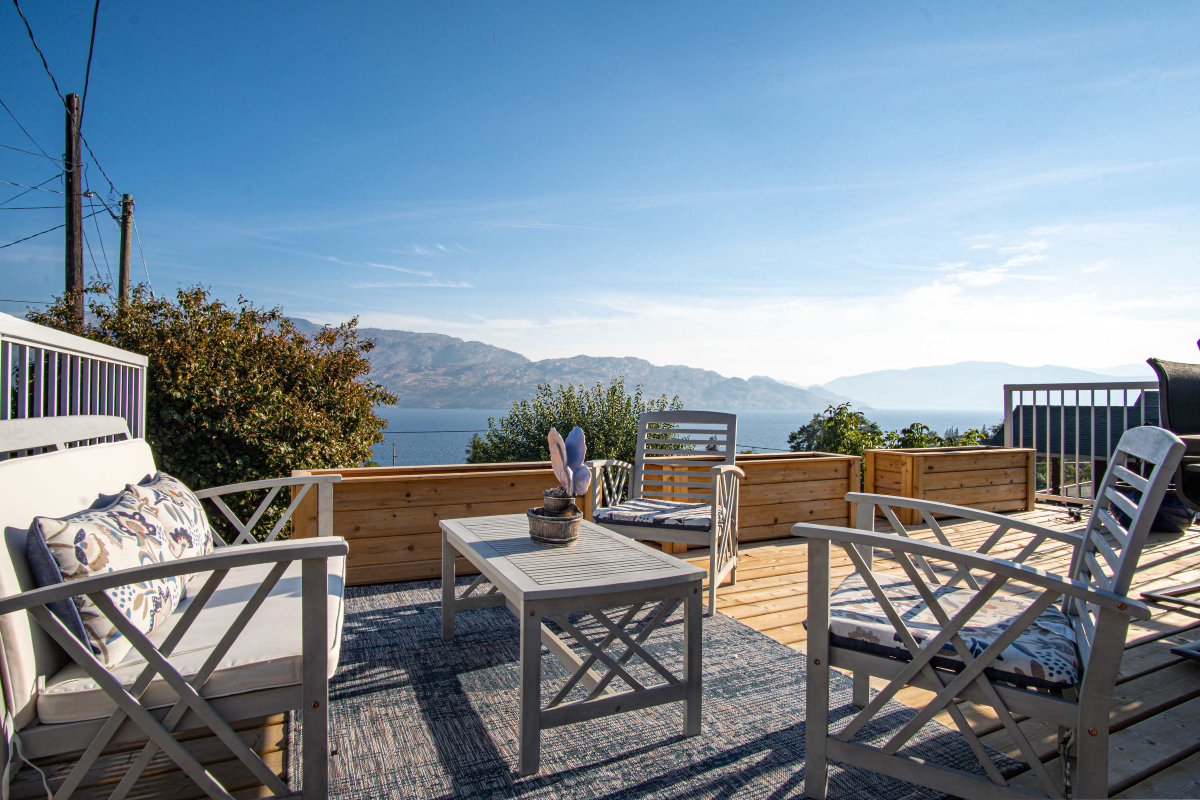 A wooden deck features white outdoor furniture, including a table and chairs, with a view of a lake surrounded by mountains.