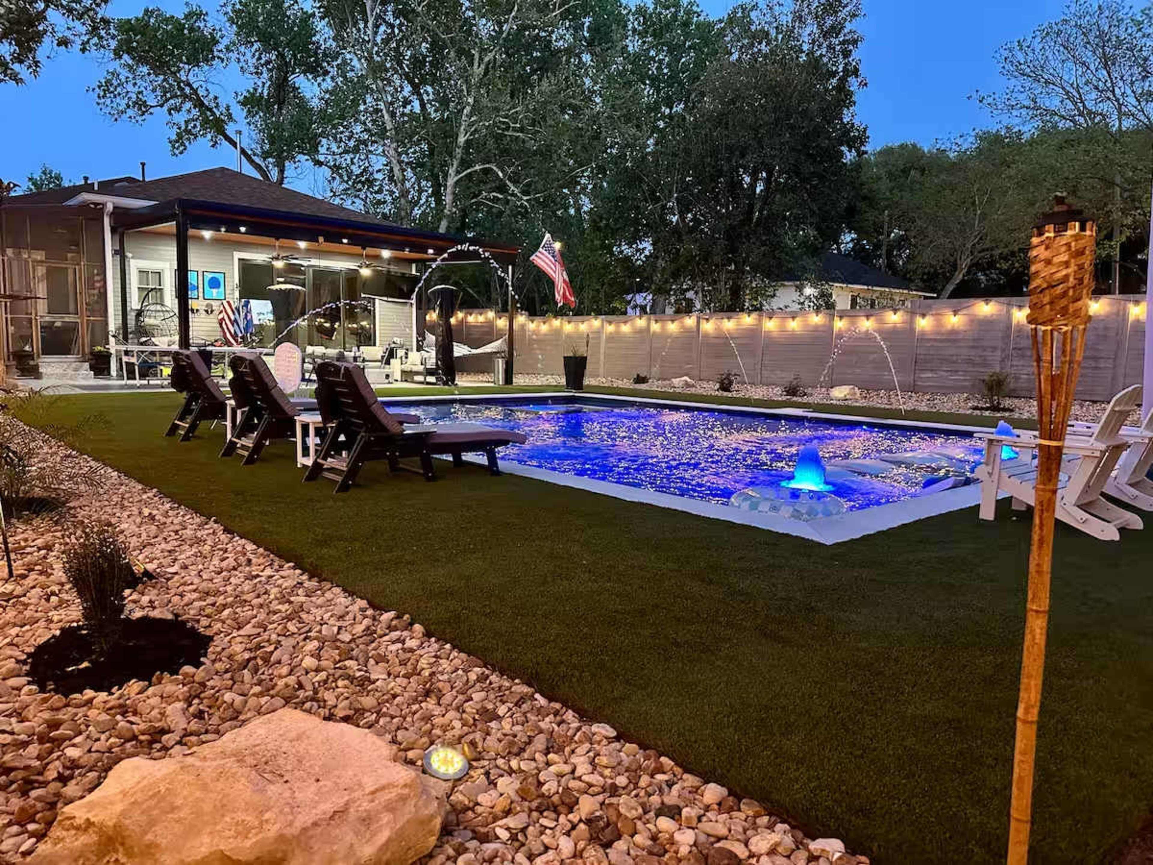 The image shows a backyard pool area featuring a lit pool surrounded by lounge chairs, artificial grass, and decorative rocks, with a house and trees in the background.