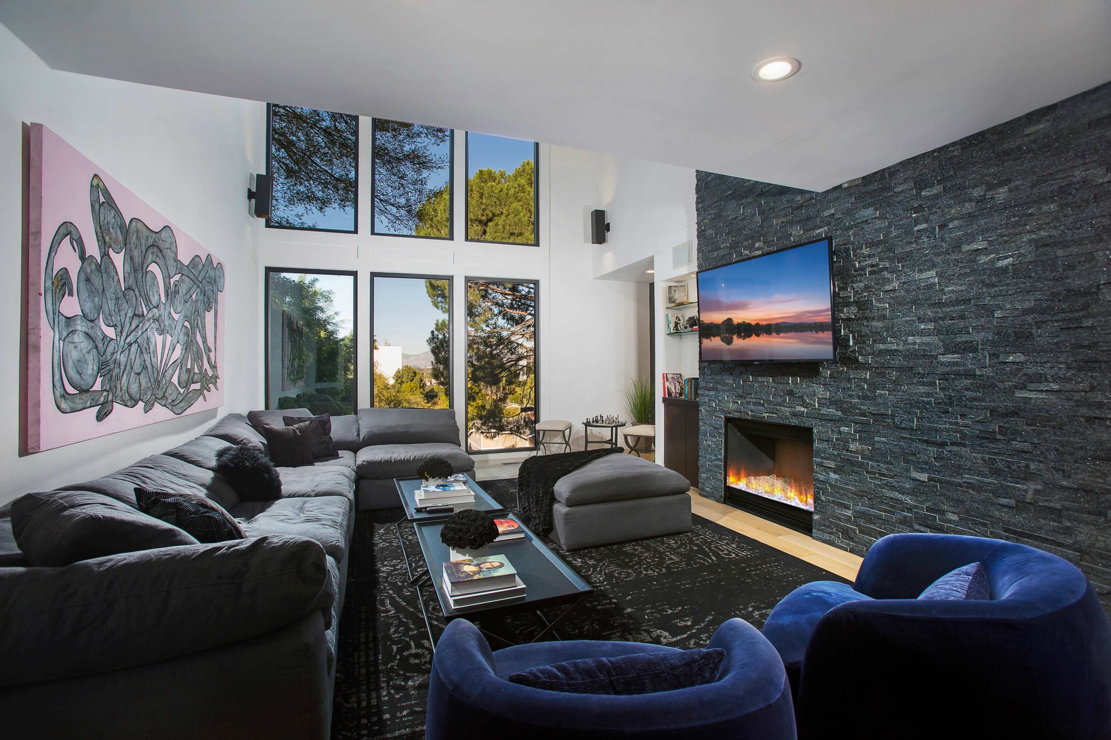 The living room features a sectional sofa, an armchair, a modern television mounted above a fireplace, and large windows showcasing an outdoor view.