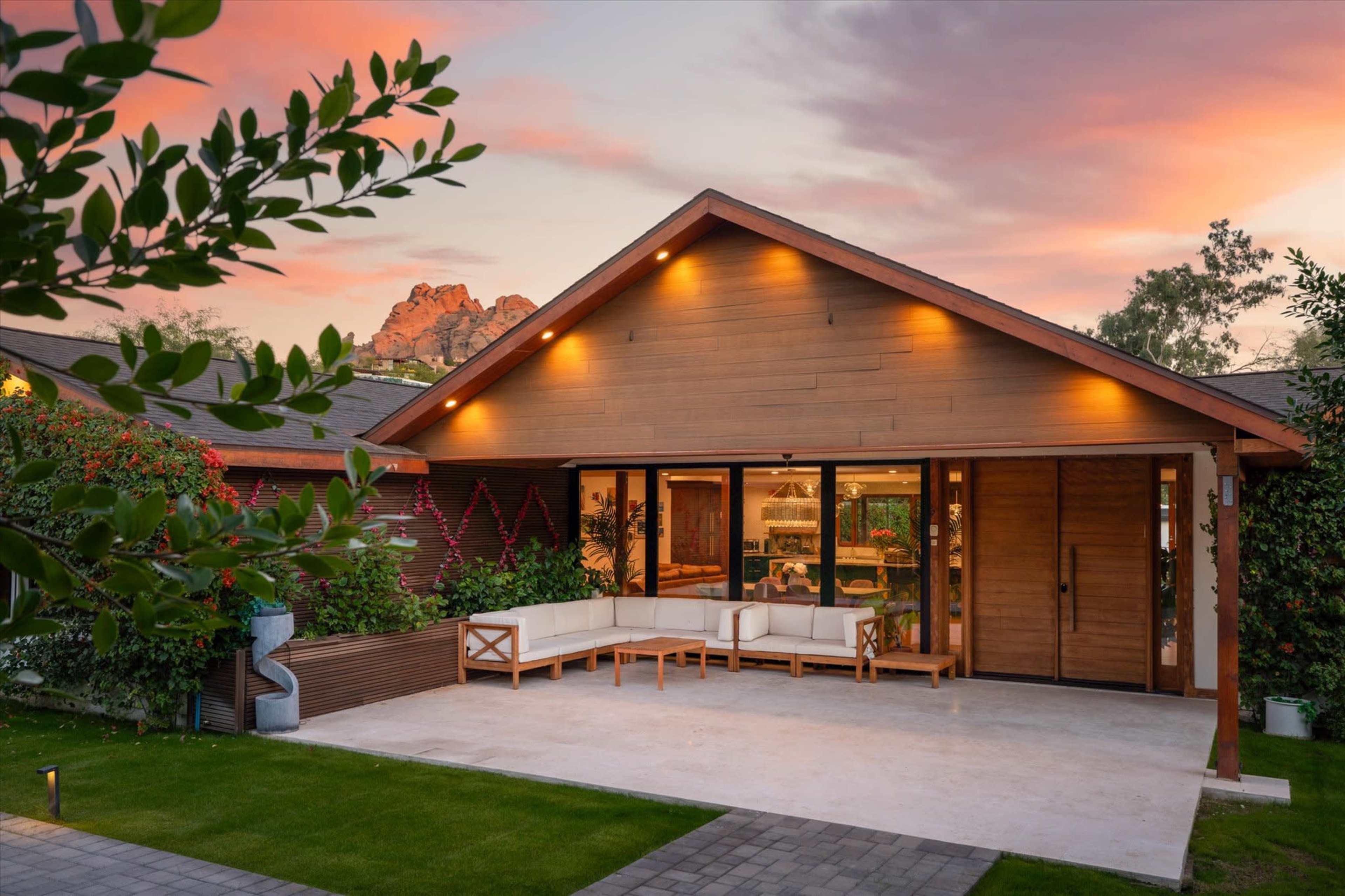 A modern wooden house features a spacious patio with white outdoor seating, surrounded by lush greenery and dramatic sunset skies.