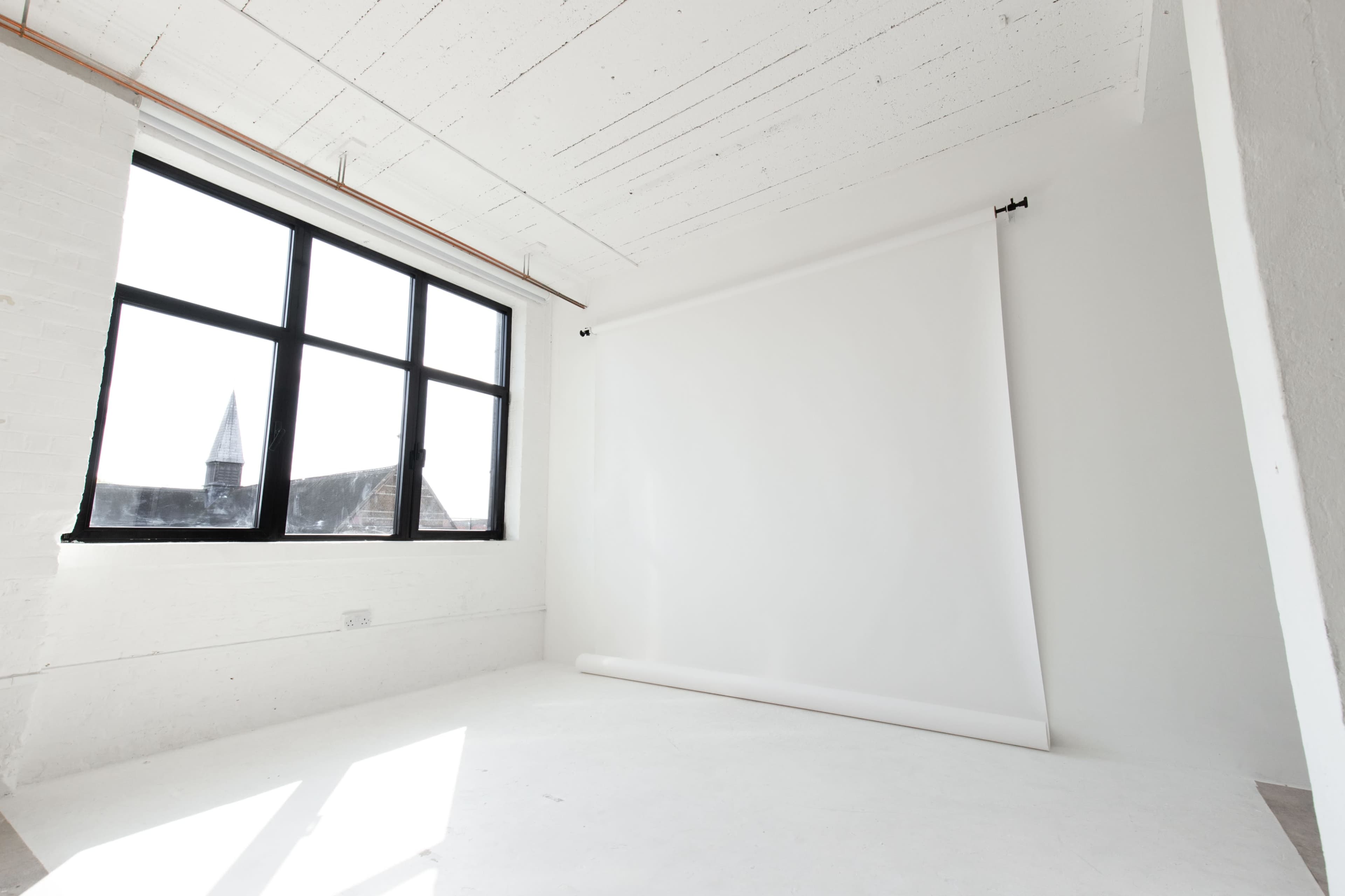 A minimalistic studio interior with a large window and a white backdrop roll against a white wall.
