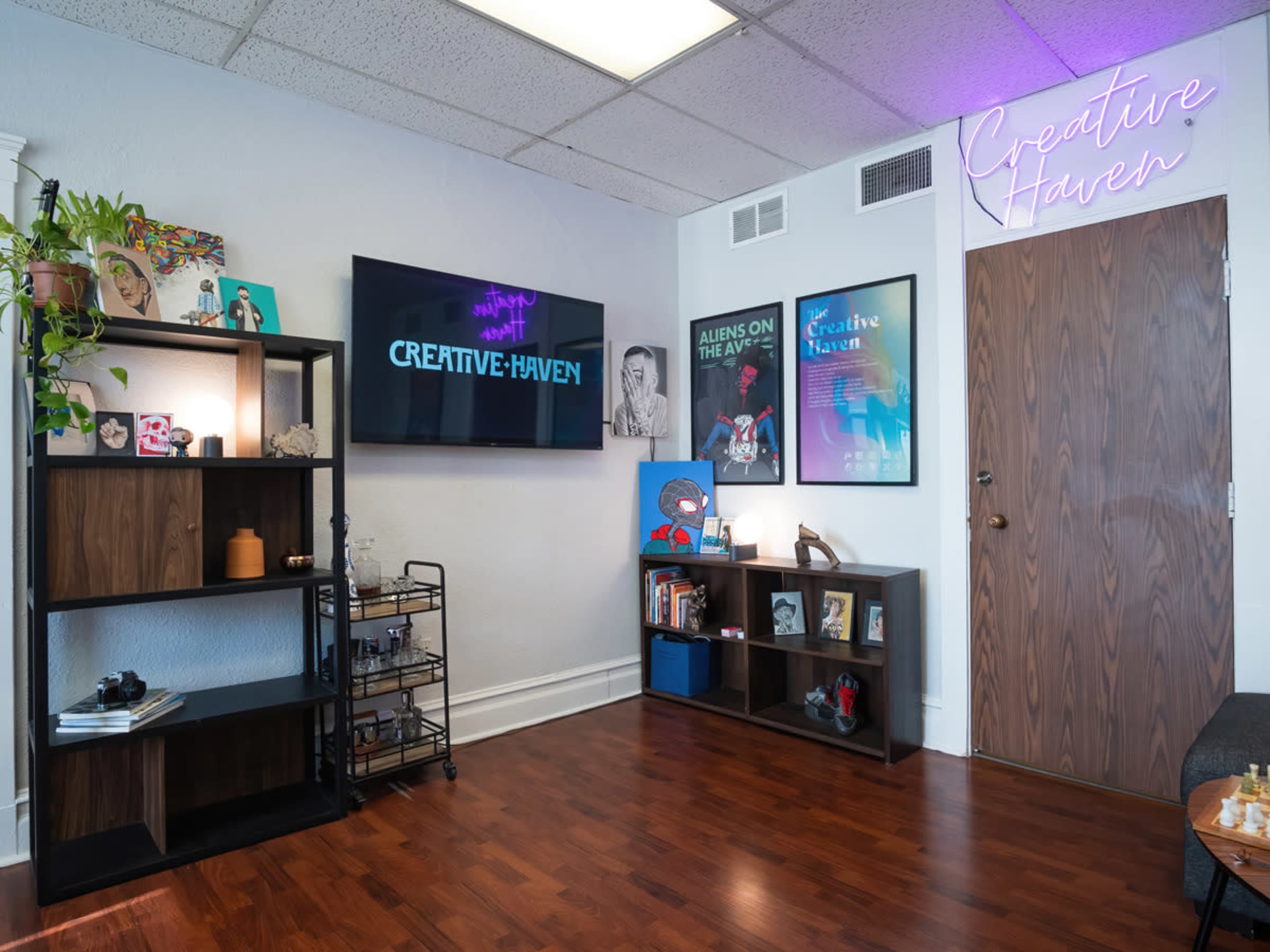 A cozy room featuring shelves with decorative items, a wall-mounted TV displaying "CREATIVE HAVEN," and a door with a neon sign above it.