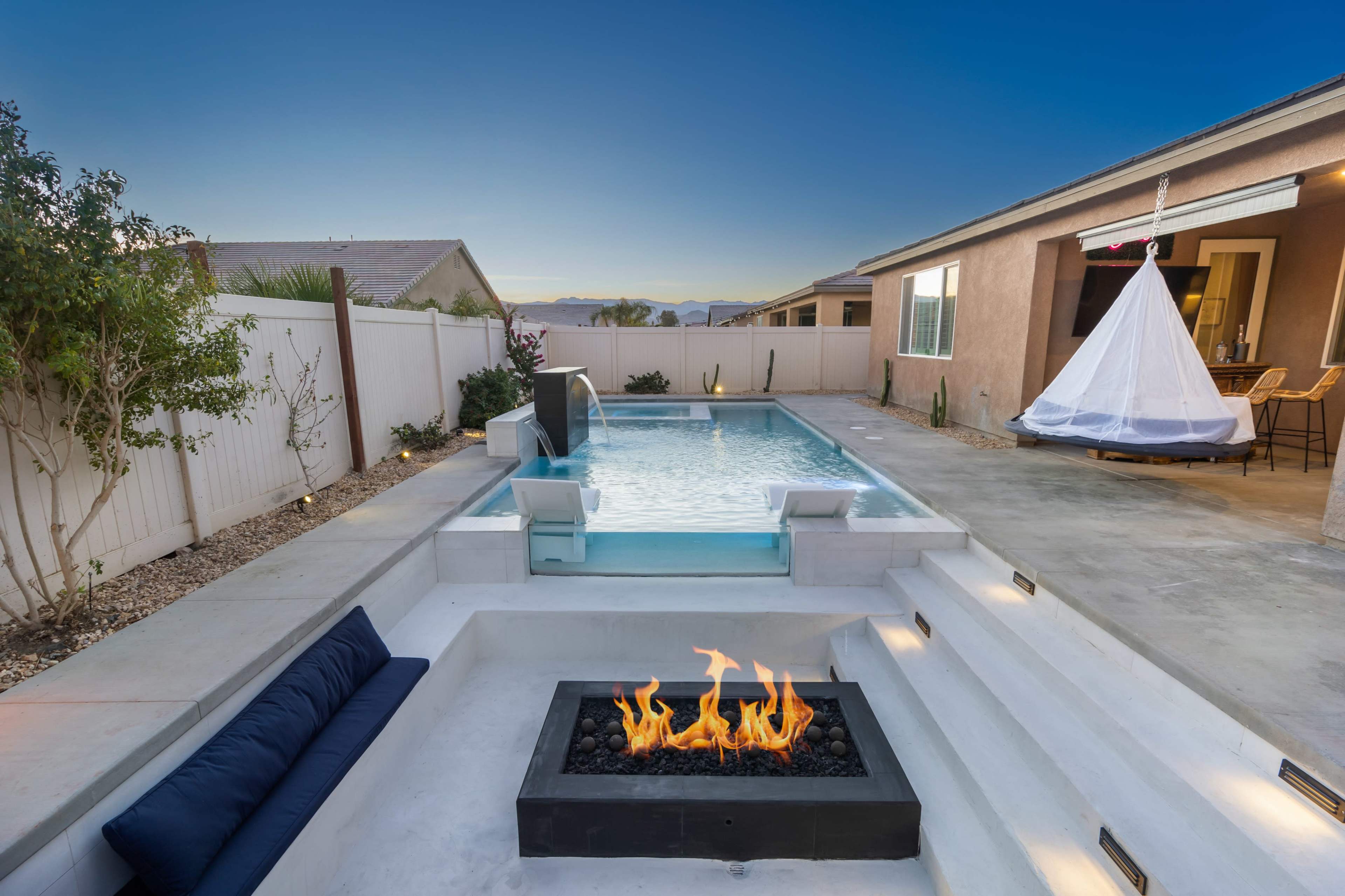 A modern backyard with a swimming pool, a fire pit, and a hanging chair, surrounded by a fence and residential buildings.