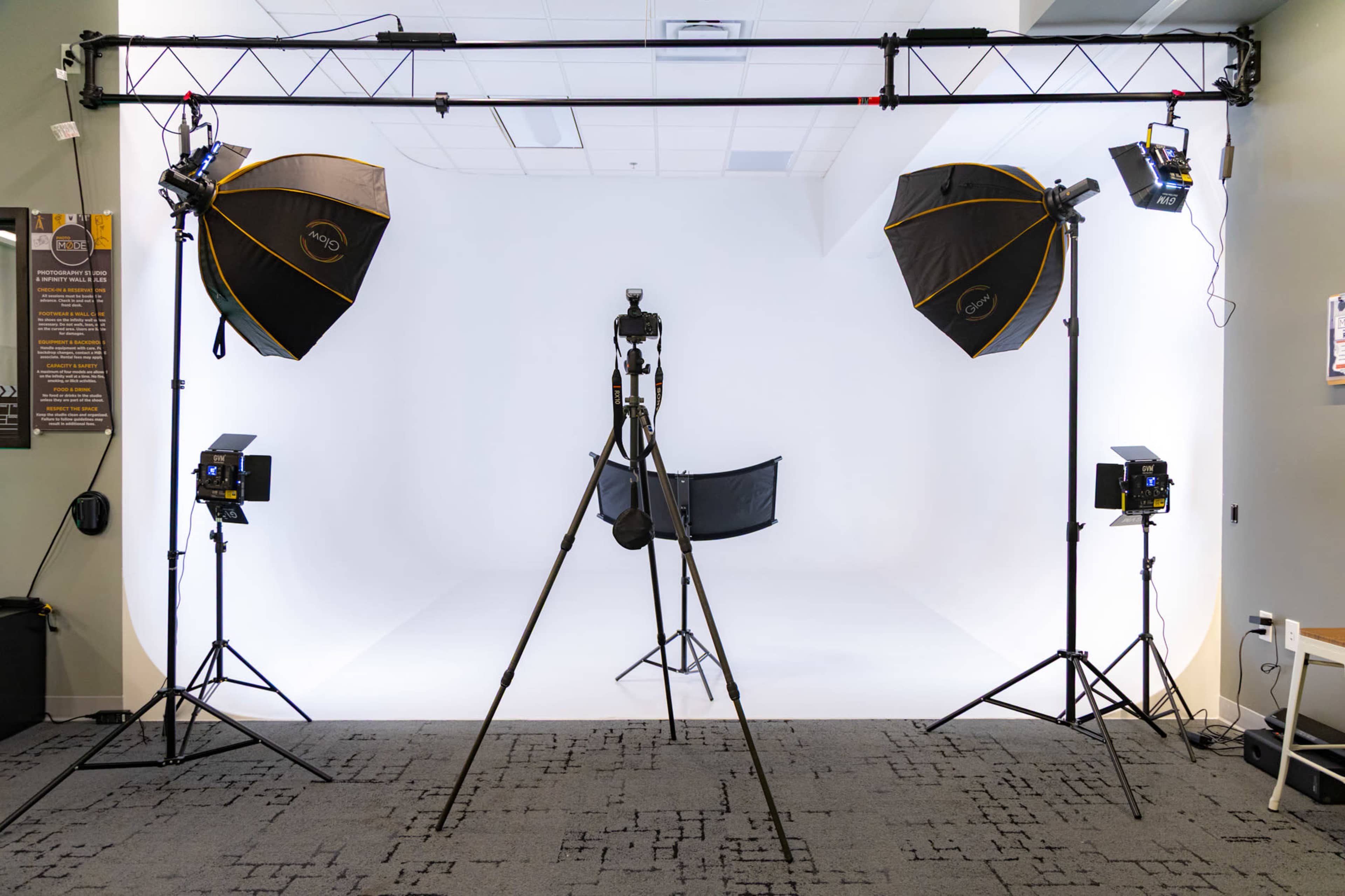 The image shows a photography studio setup with two large softbox lights, a camera on a tripod, and a backdrop on a seamless white floor.