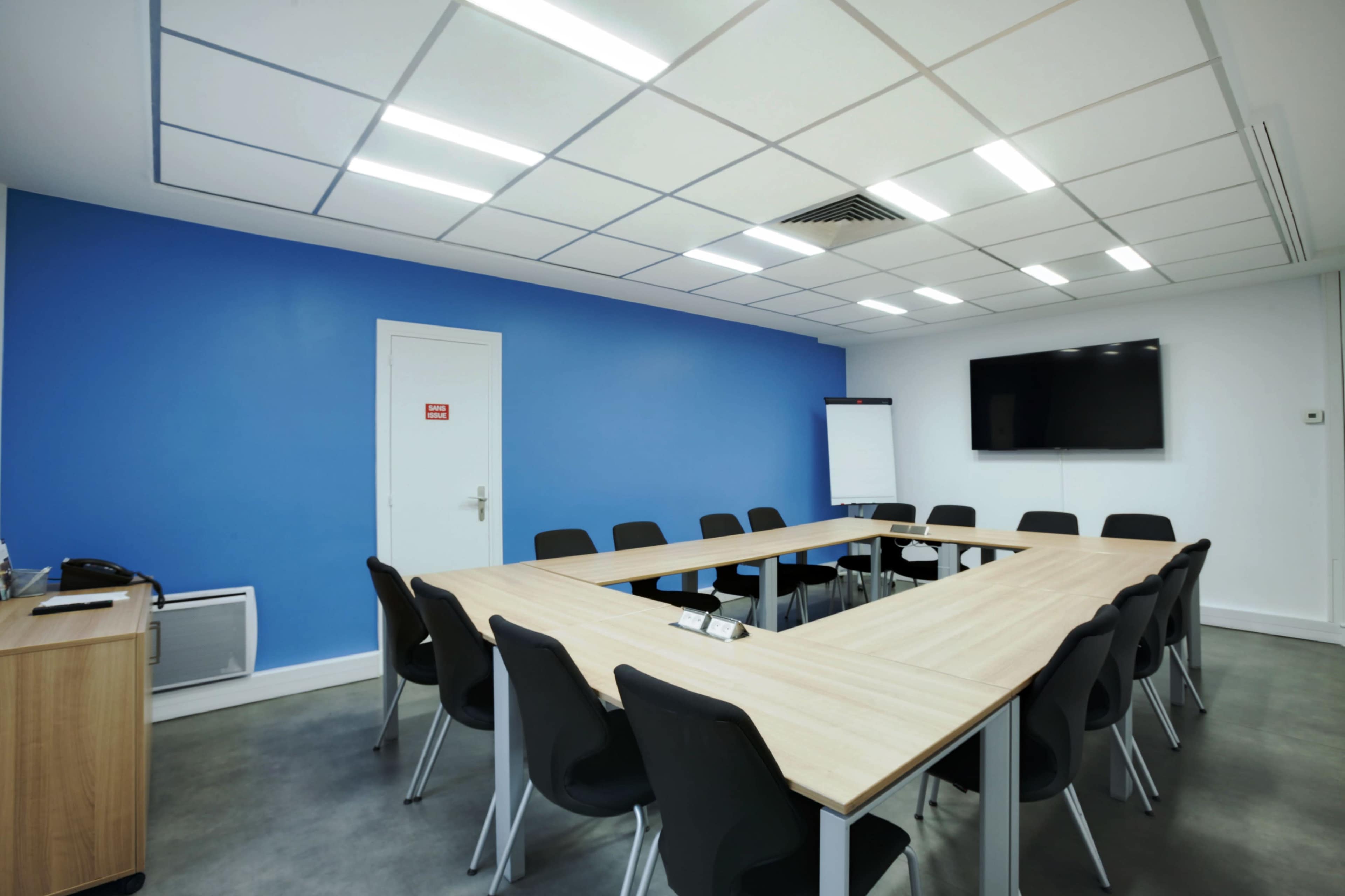 A modern conference room featuring a large wooden table arranged in a U-shape, surrounded by black chairs, with a blue accent wall and a wall-mounted TV.