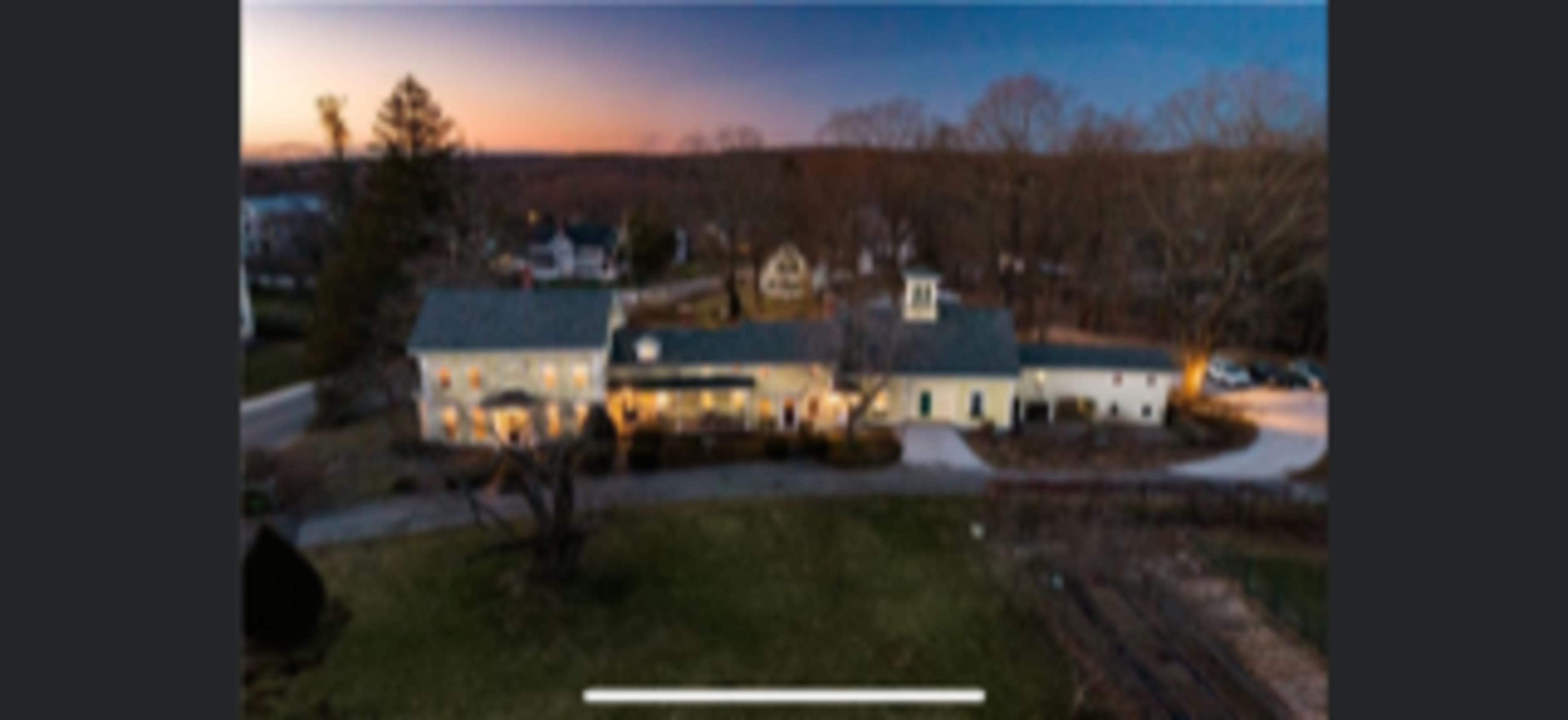 A large house with a green roof, surrounded by a spacious yard and several trees, at dusk with lights illuminated in the windows.