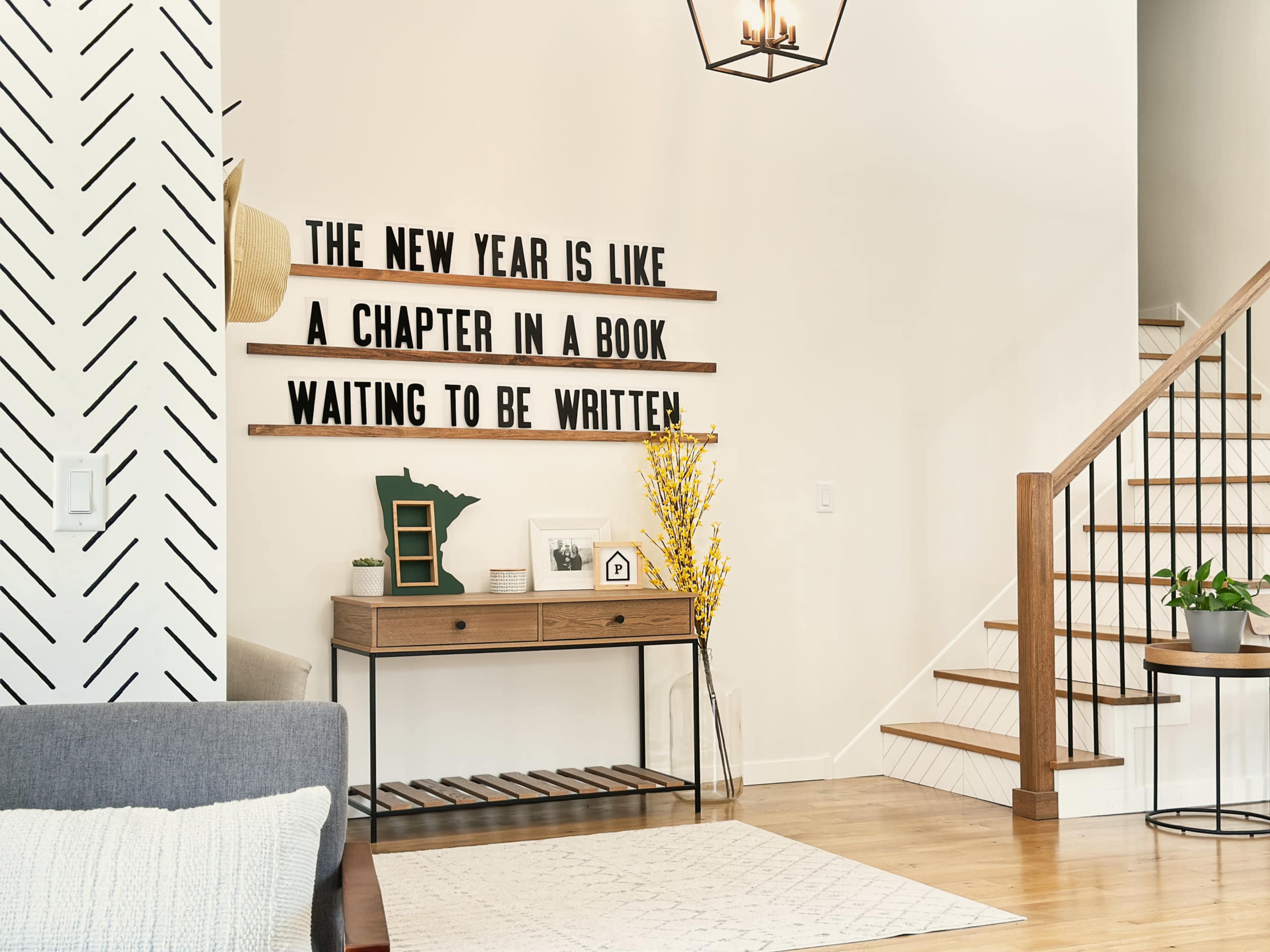 The image shows a modern living room with a decorative wall featuring wooden shelves that display the phrase "THE NEW YEAR IS LIKE A CHAPTER IN A BOOK WAITING TO BE WRITTEN" alongside a staircase and a tabletop decor setup.