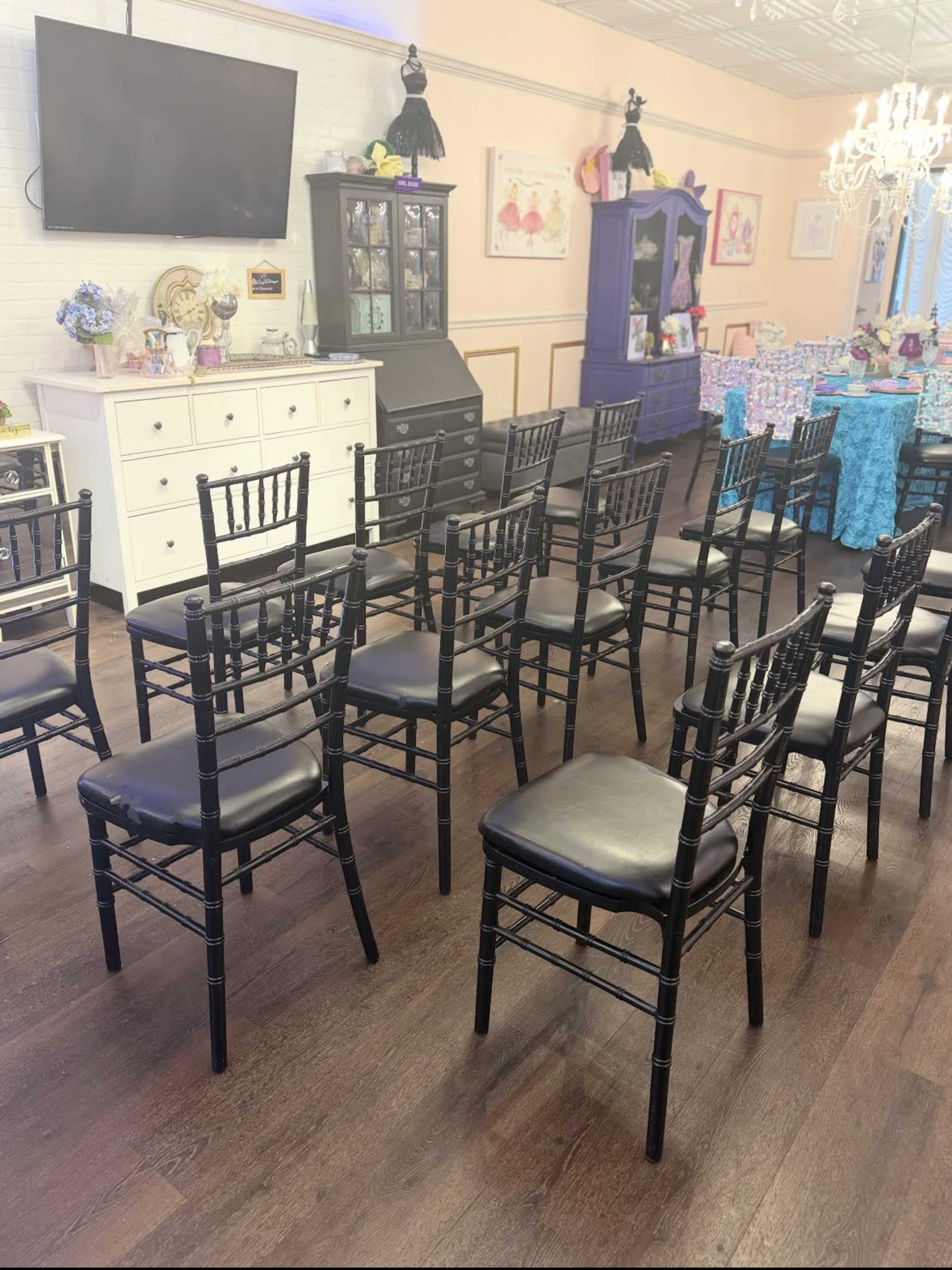 A room with black chairs arranged in a circular formation around a central space, featuring a television and various pieces of furniture in the background.