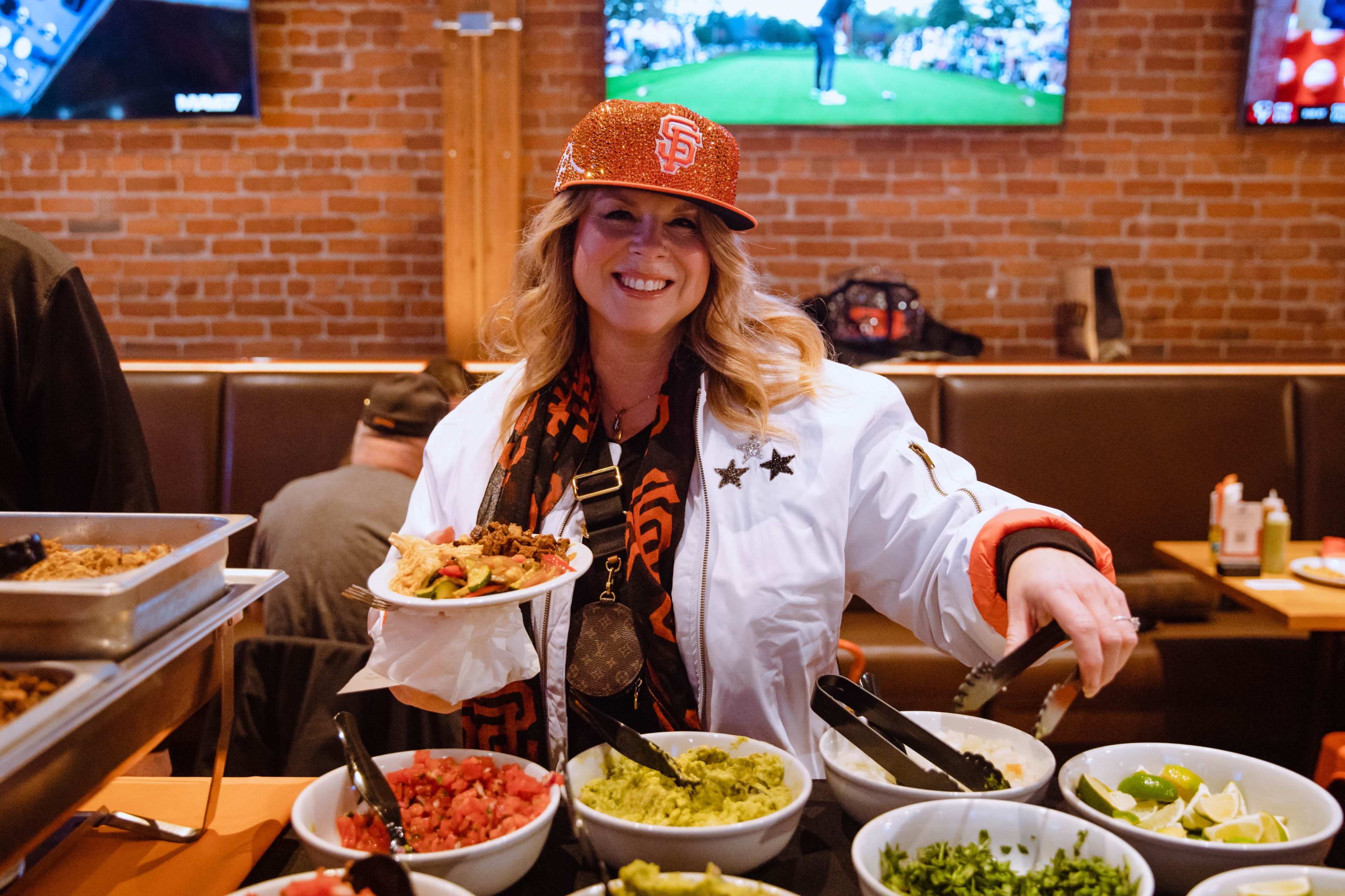 A woman in a San Francisco Giants cap and jacket smiles while serving food from a buffet table featuring various toppings like guacamole, salsa, and diced tomatoes.