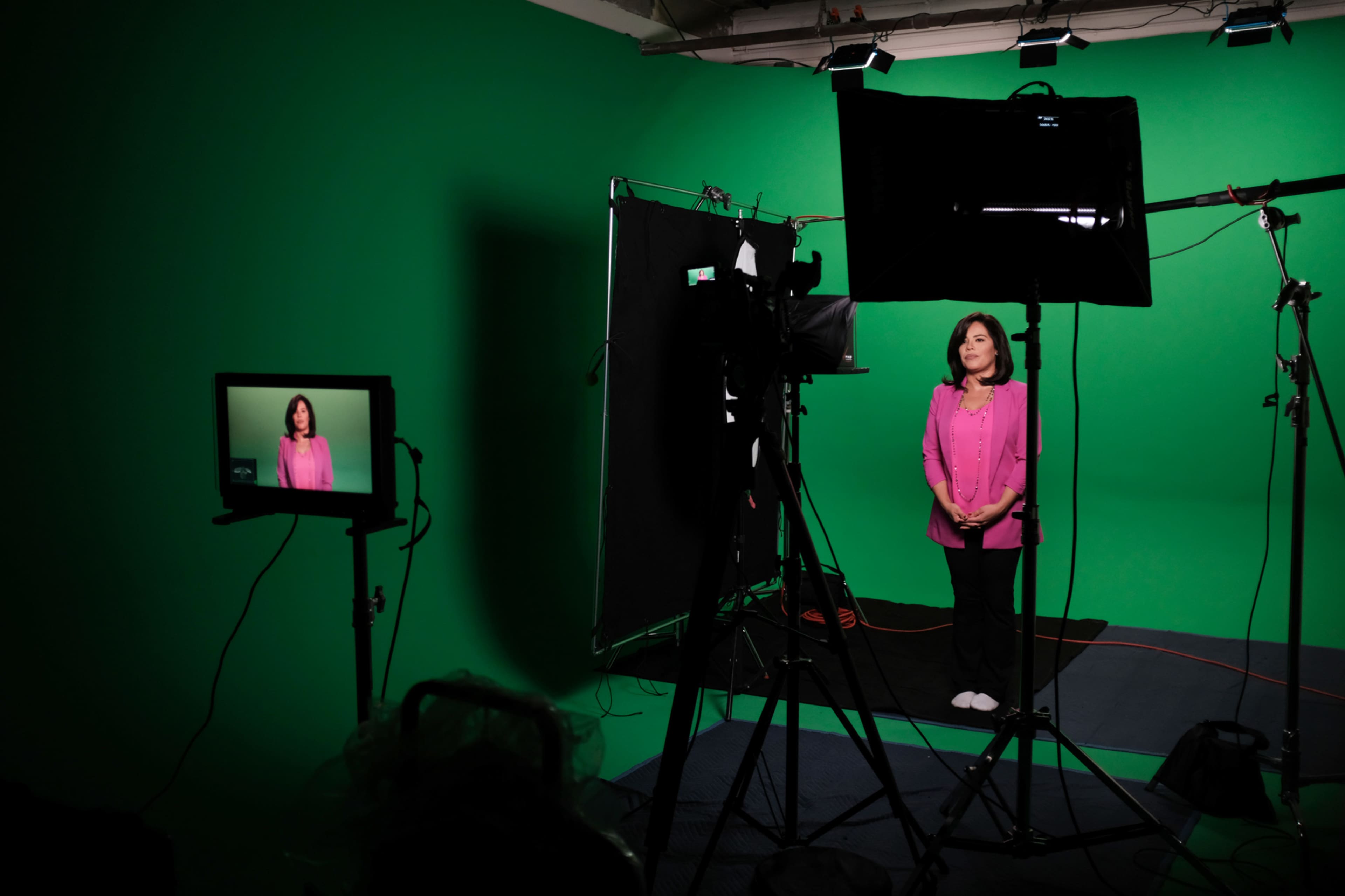A woman in a pink top stands in front of a green screen while being filmed, with camera equipment and a monitor positioned nearby.