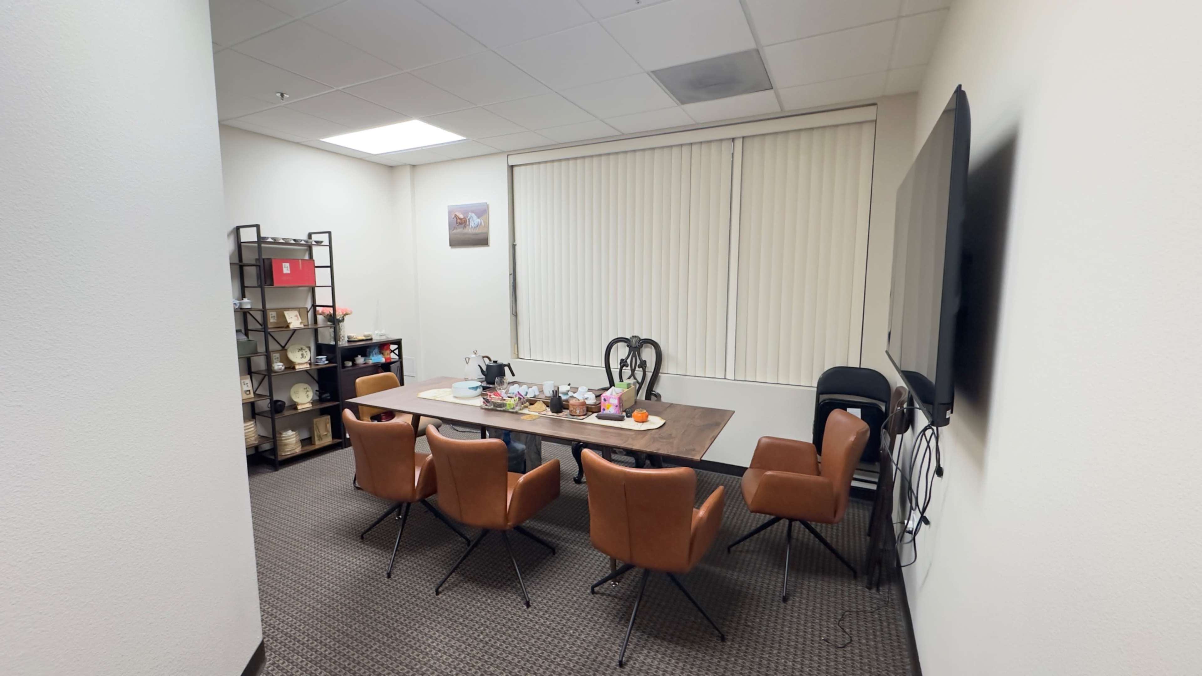 The image shows a well-lit conference room with a large table surrounded by six brown chairs, a shelf filled with various items, and a television mounted on the wall.