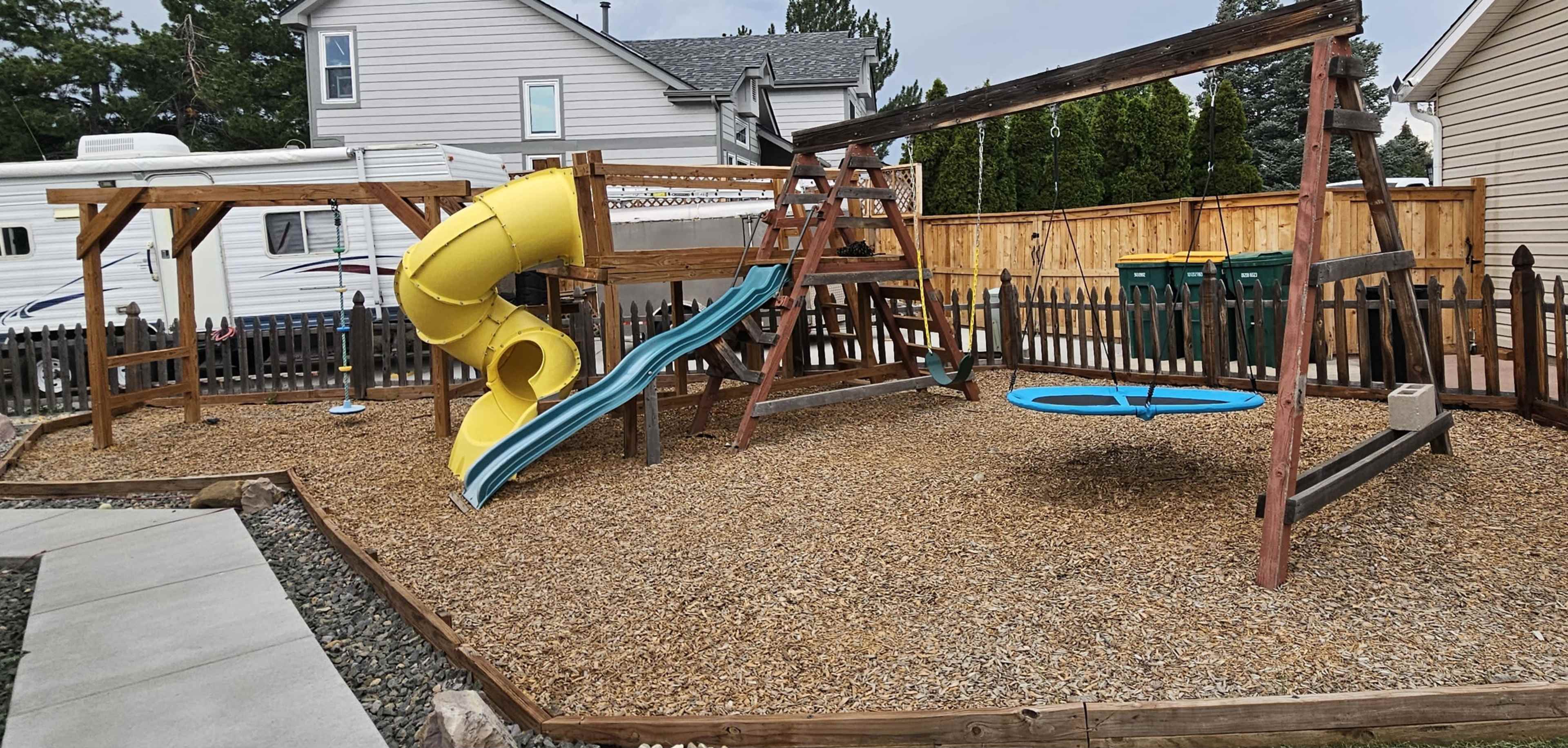 The image shows a playground with a yellow slide, a climbing structure, and a swing set, all surrounded by a gravel surface and a wooden fence.