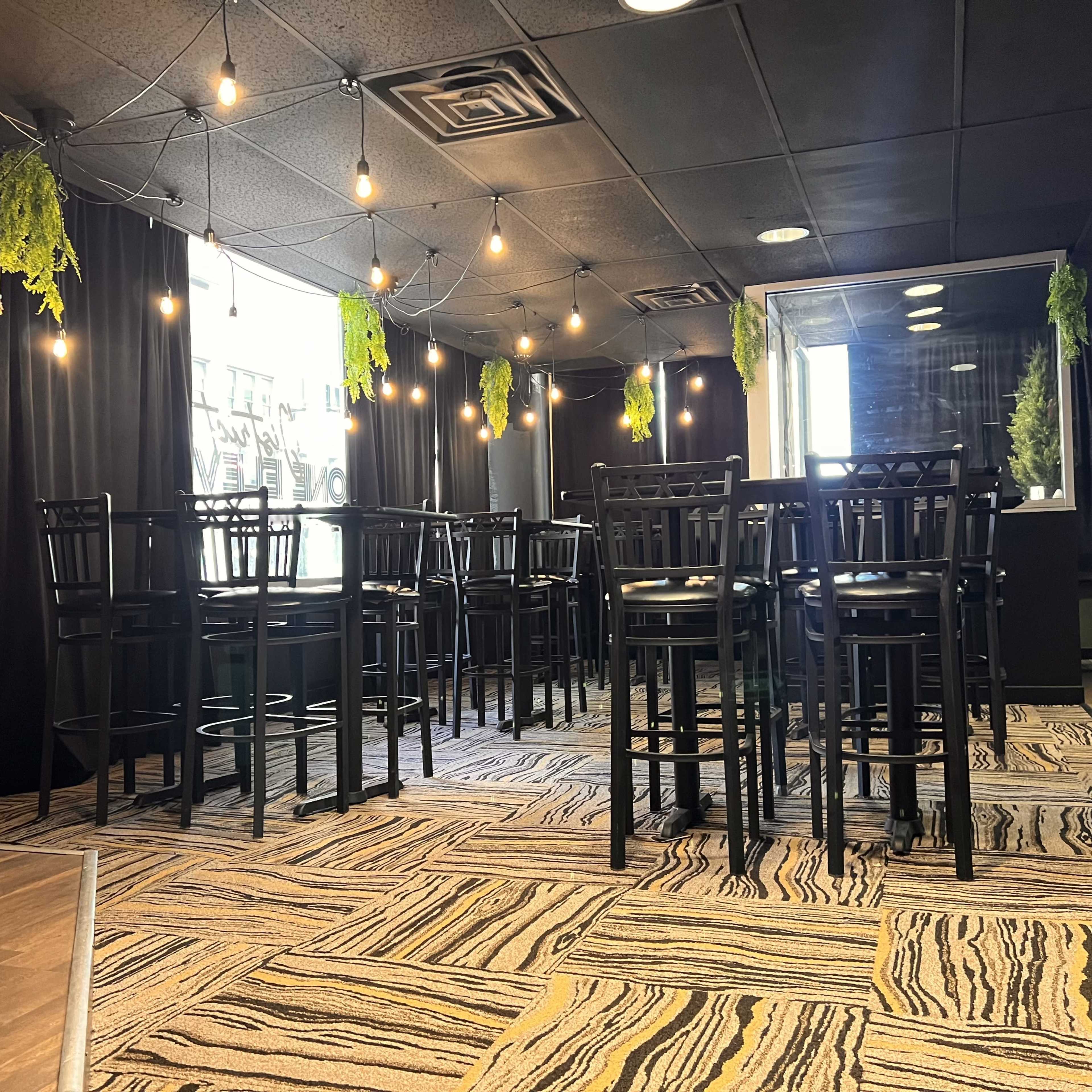 The image shows a room with several high-top black chairs and tables, illuminated by string lights and adorned with green plants.