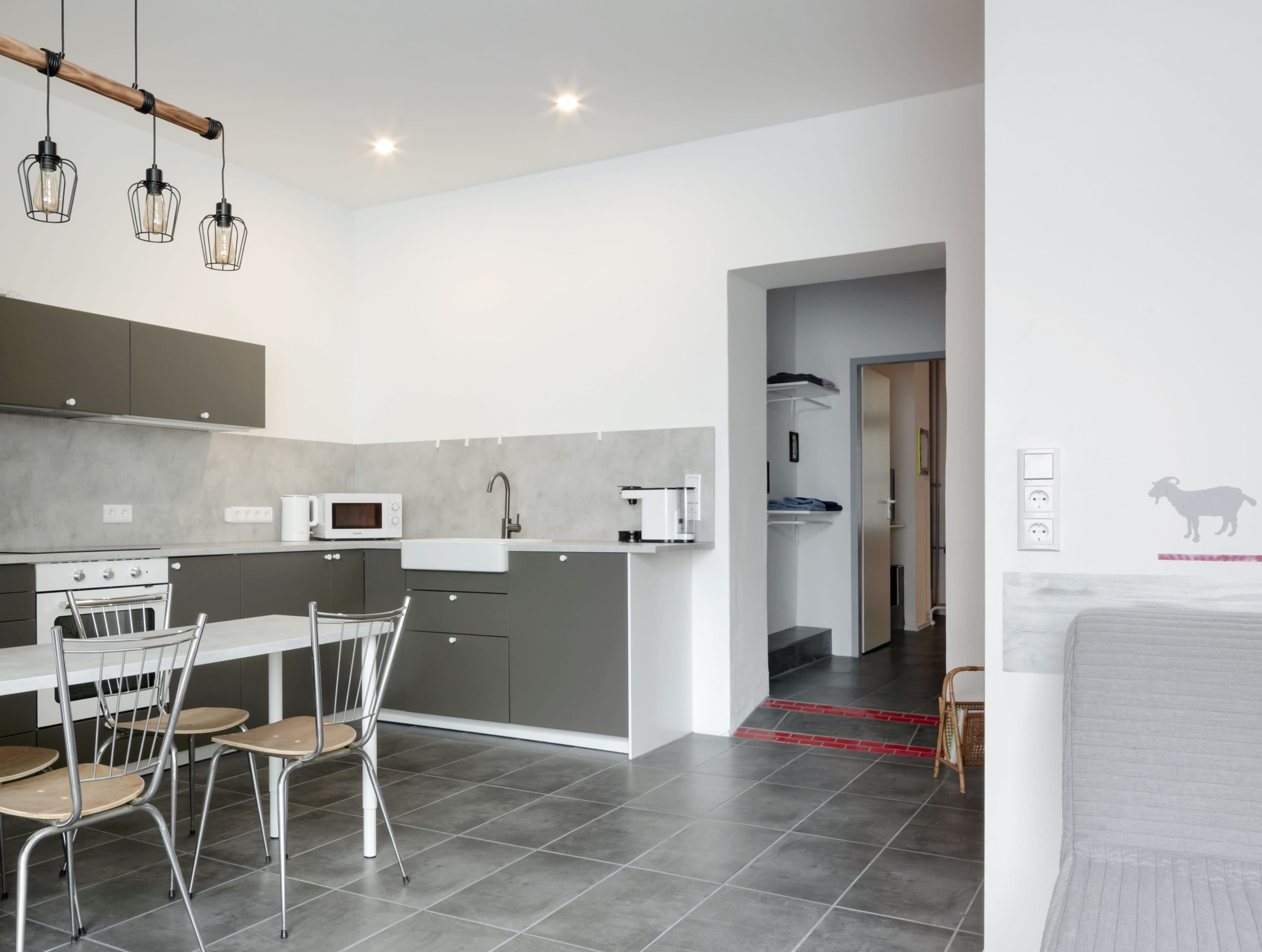 The image shows a modern kitchen with gray cabinets, a white countertop, and a dining table with metal chairs, along with an entrance to another room in the background.