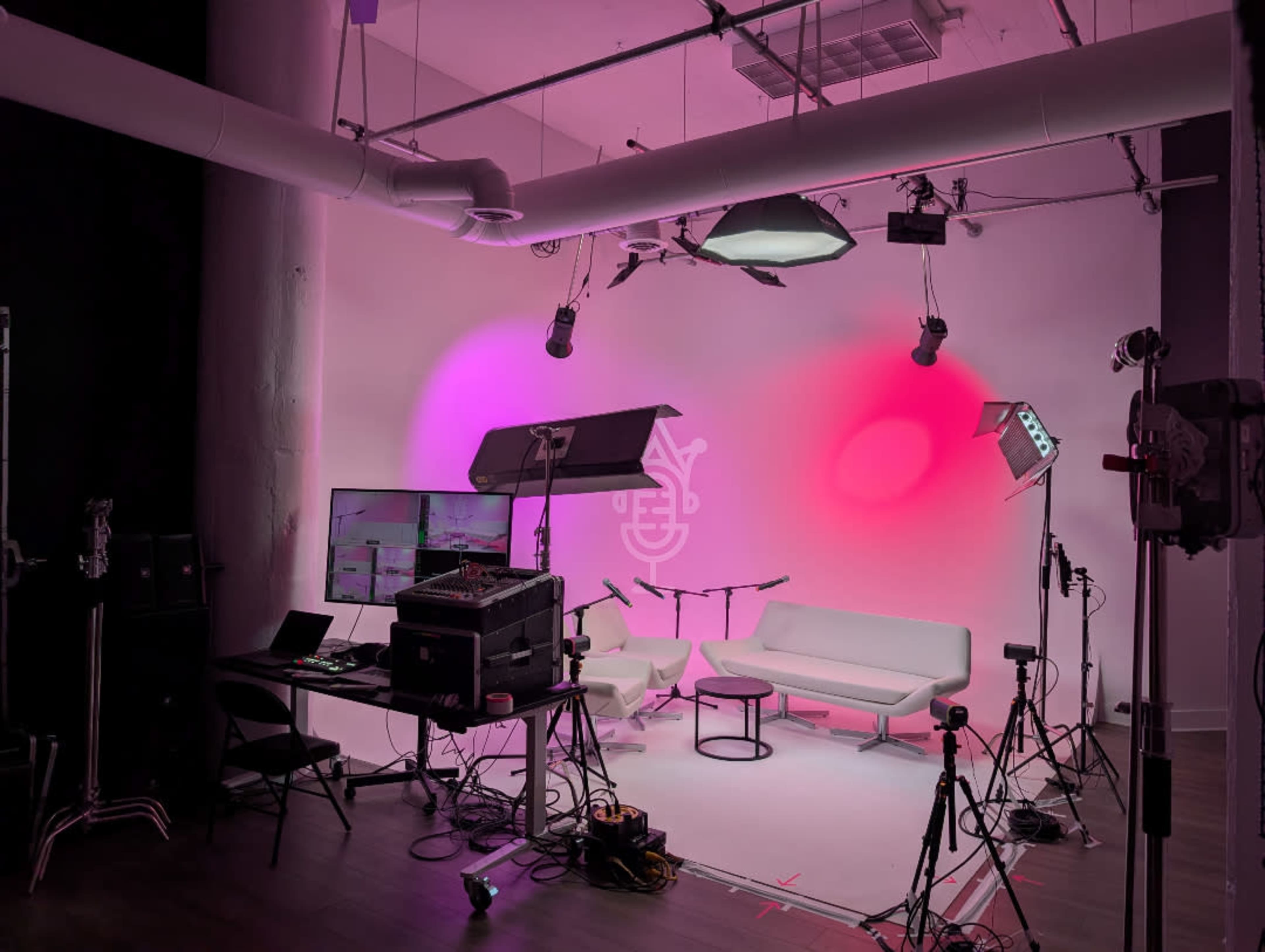 The image shows a studio setup with pink and purple lighting, two white couches, a television monitor, and various filming equipment.