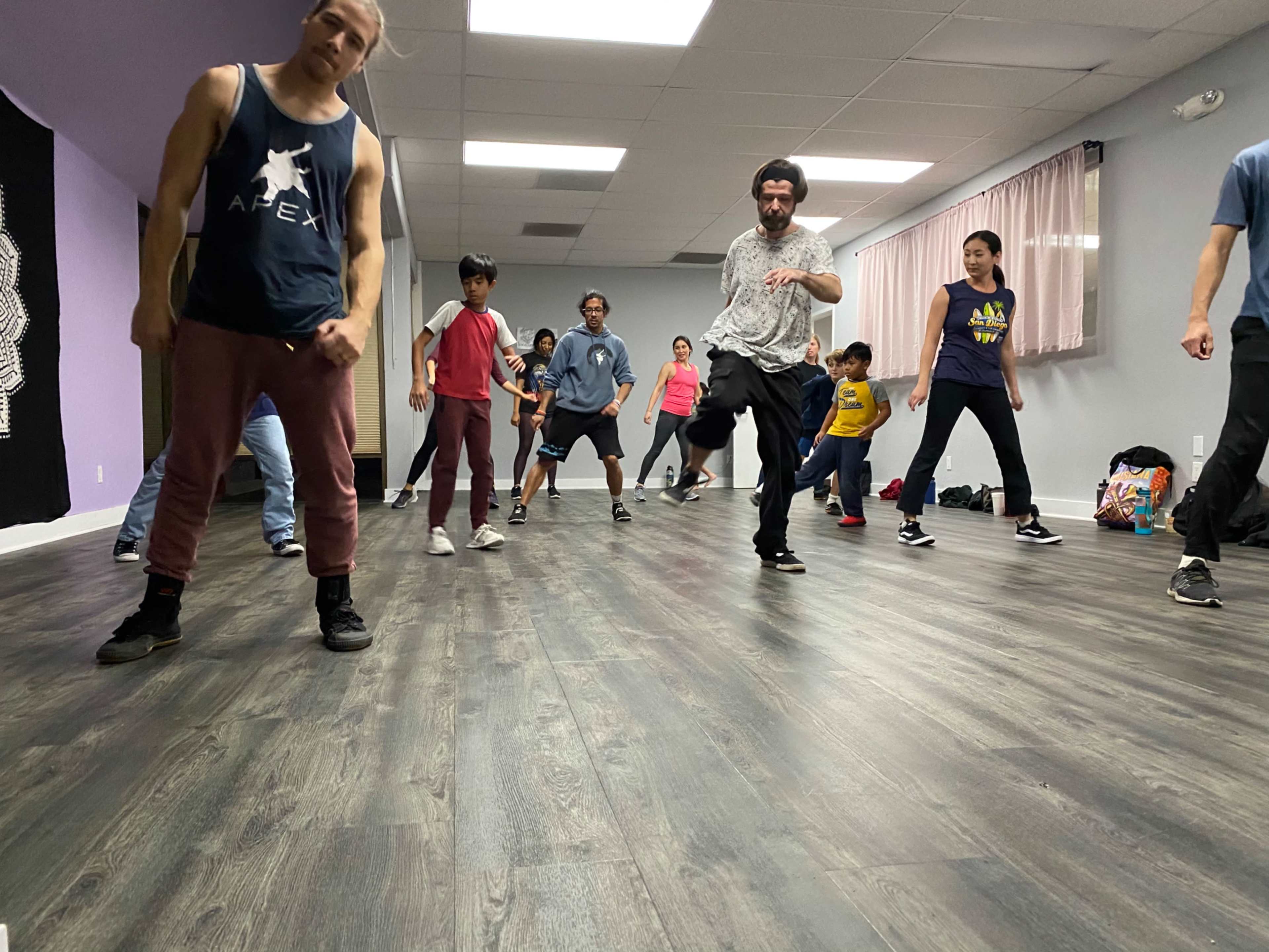 A group of people dance in a studio with wooden floors and mirrored walls.
