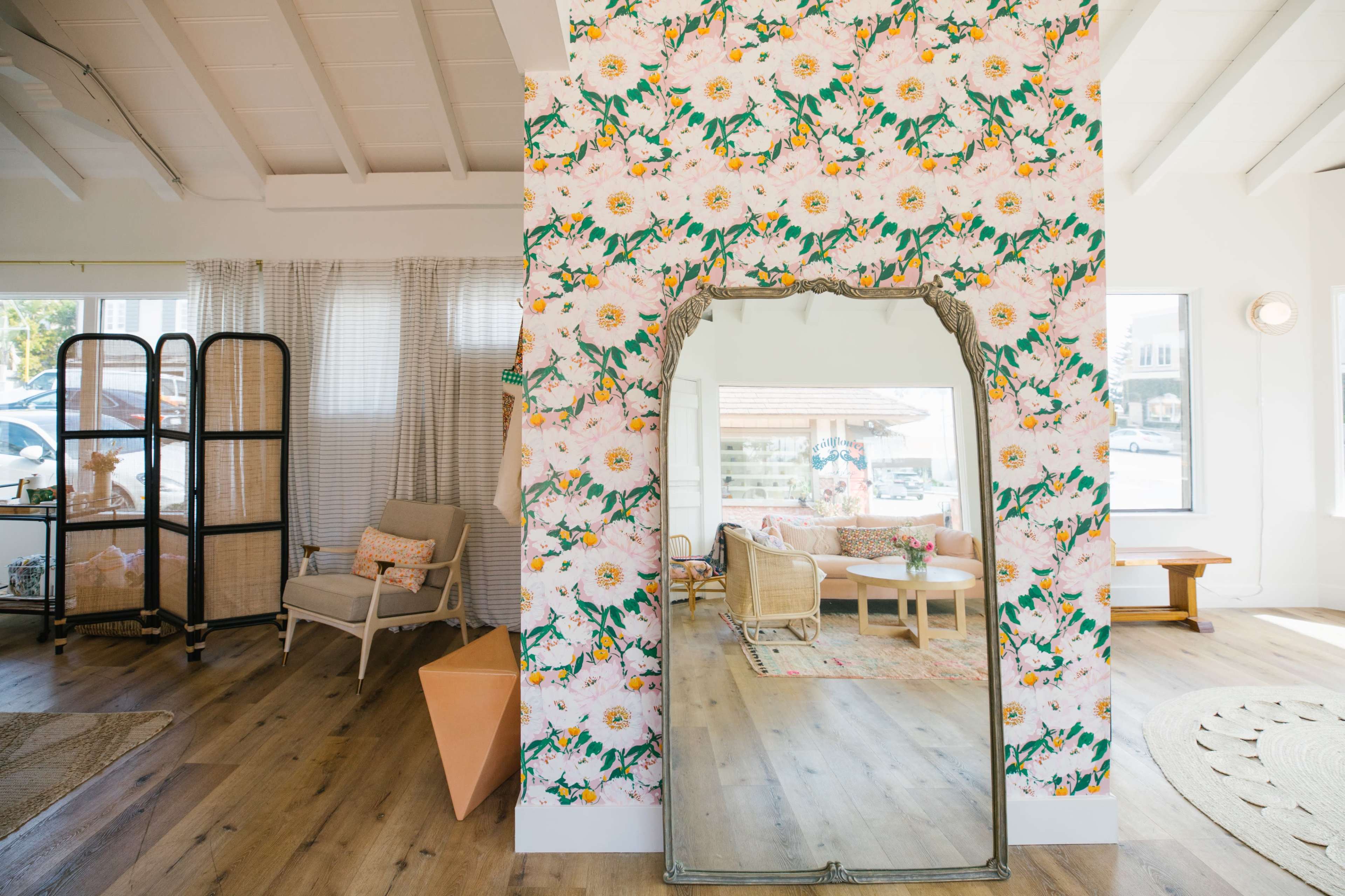 A bright, open living space features a floral-patterned wall, a large mirror, and a cozy seating area with natural light filtering through the windows.