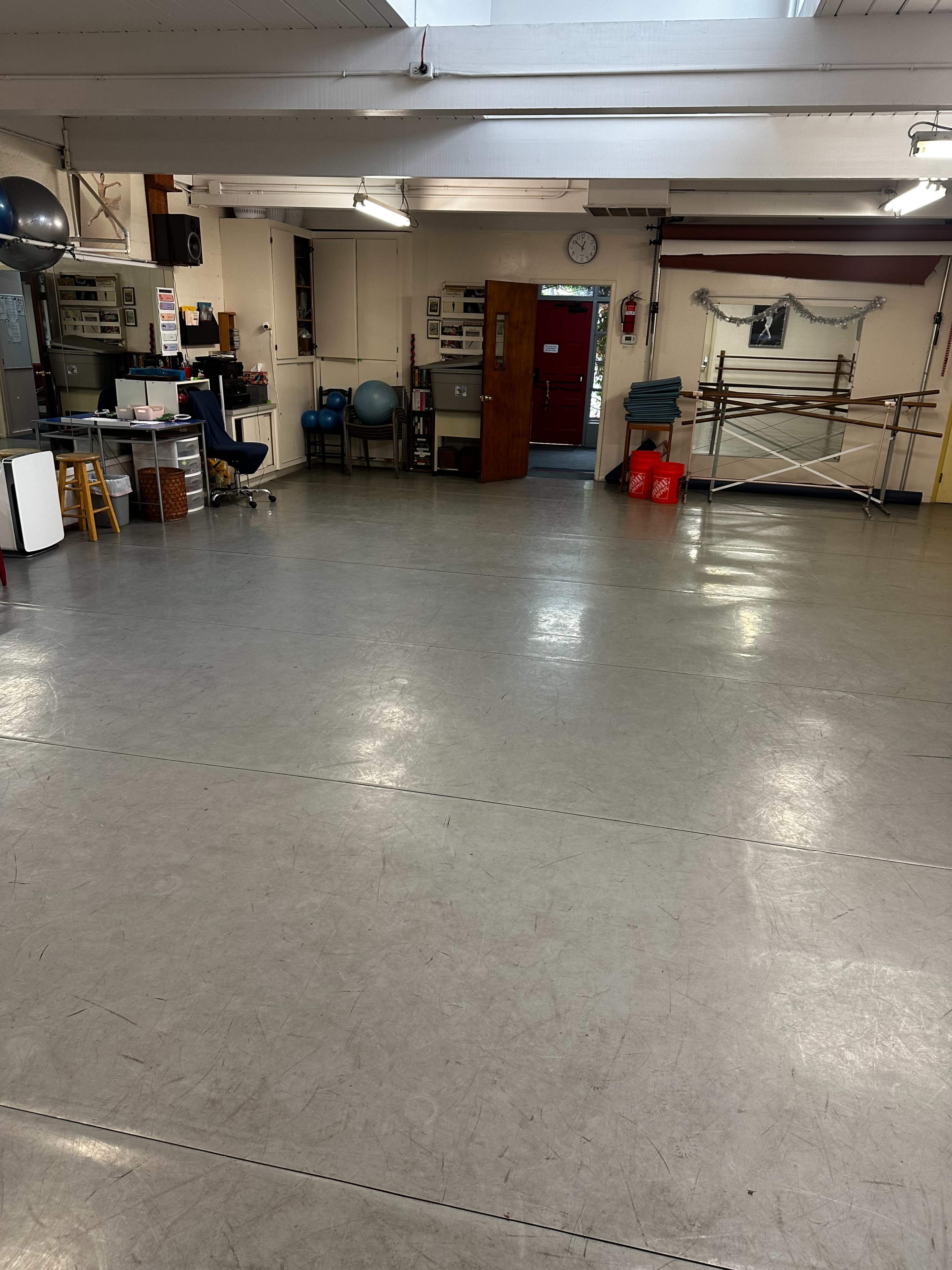 The image shows an empty studio space with a wooden barre along one wall, a desk with office equipment, and exercise equipment in the background.