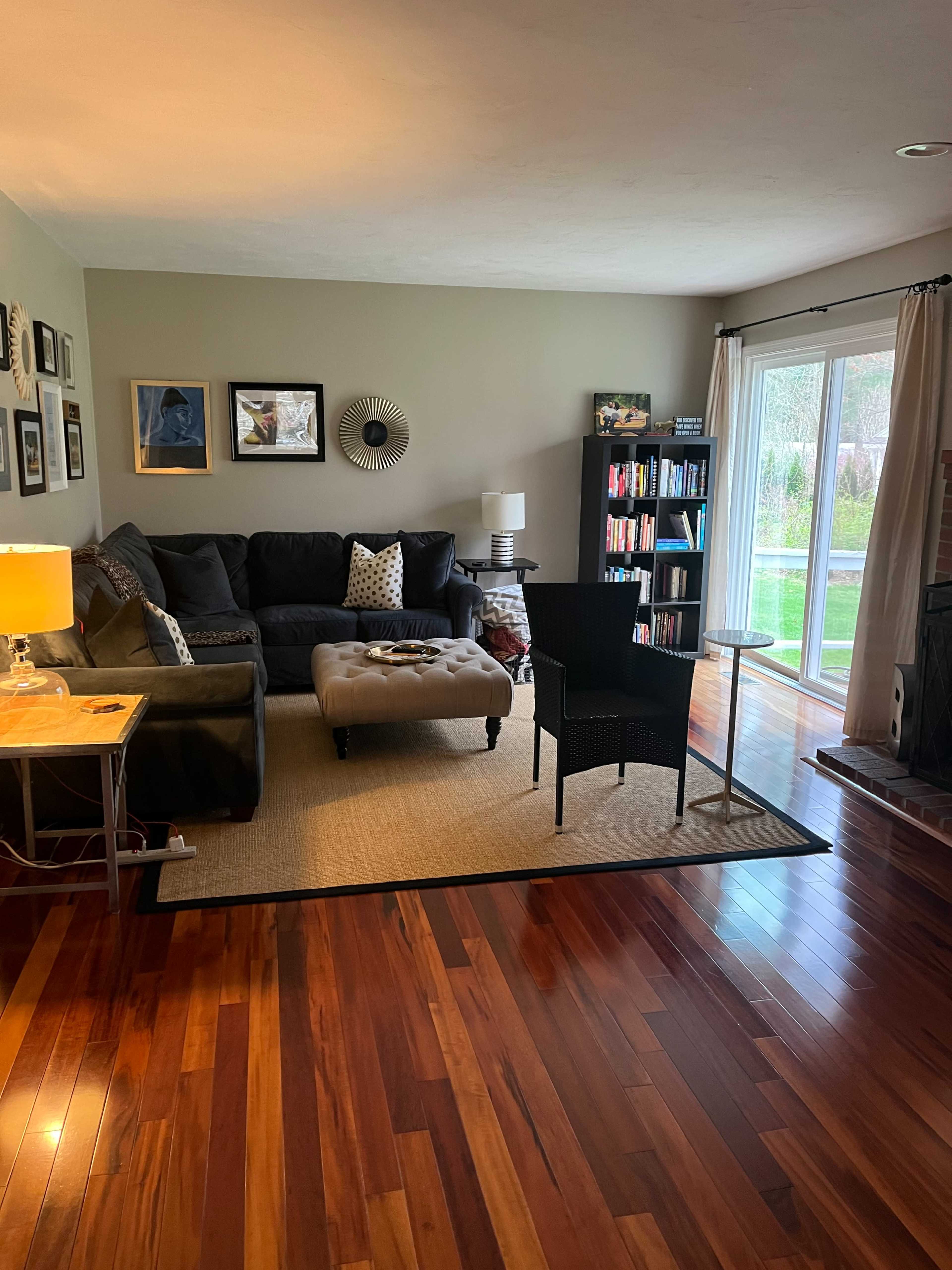 The living room features a dark sofa, an ottoman, a small table with a lamp, two chairs, and a bookshelf, all set against wooden flooring and large windows overlooking a garden.