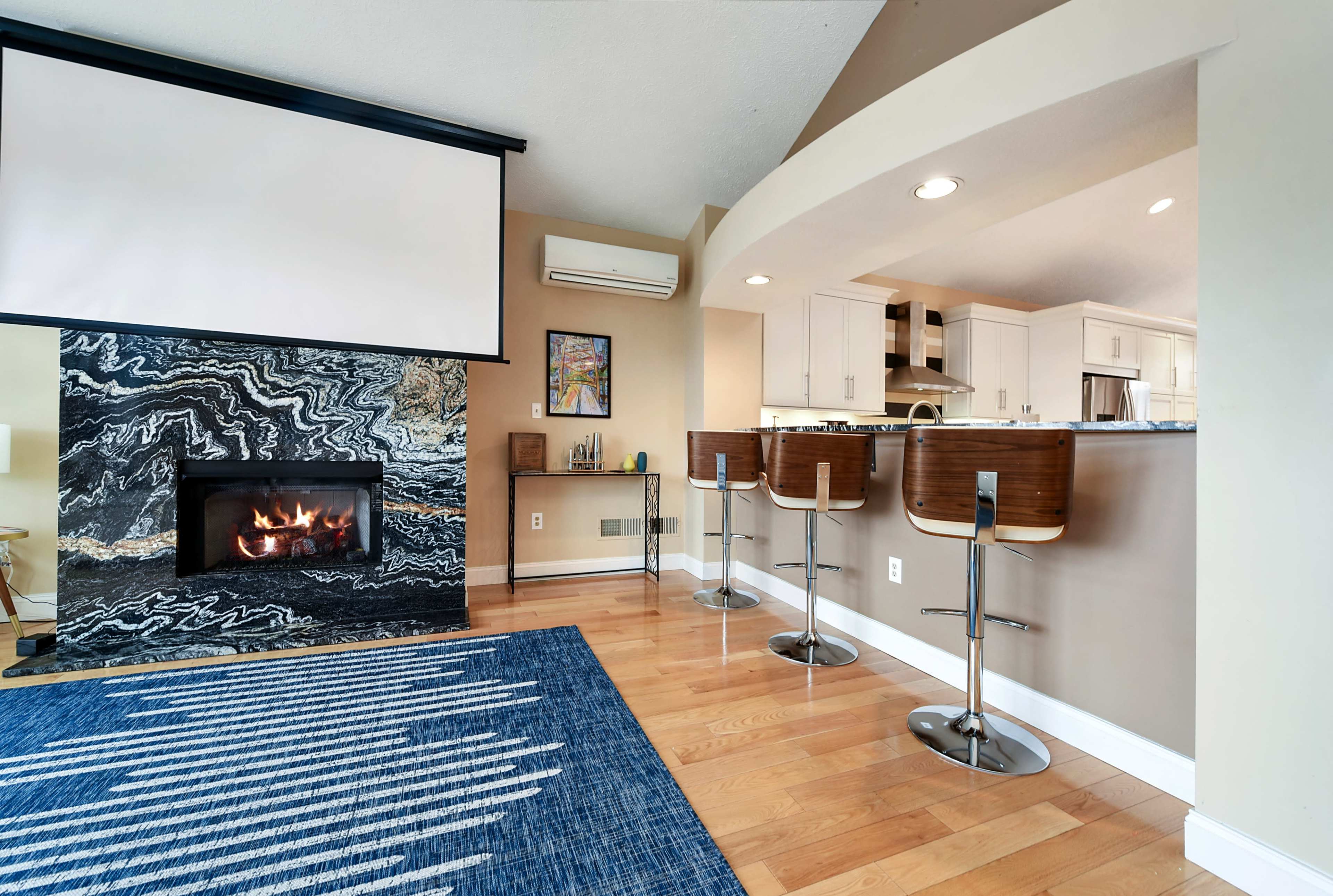A modern living area featuring a dark marble fireplace, a flat-screen projector screen, a kitchen bar with four stools, and hardwood flooring.
