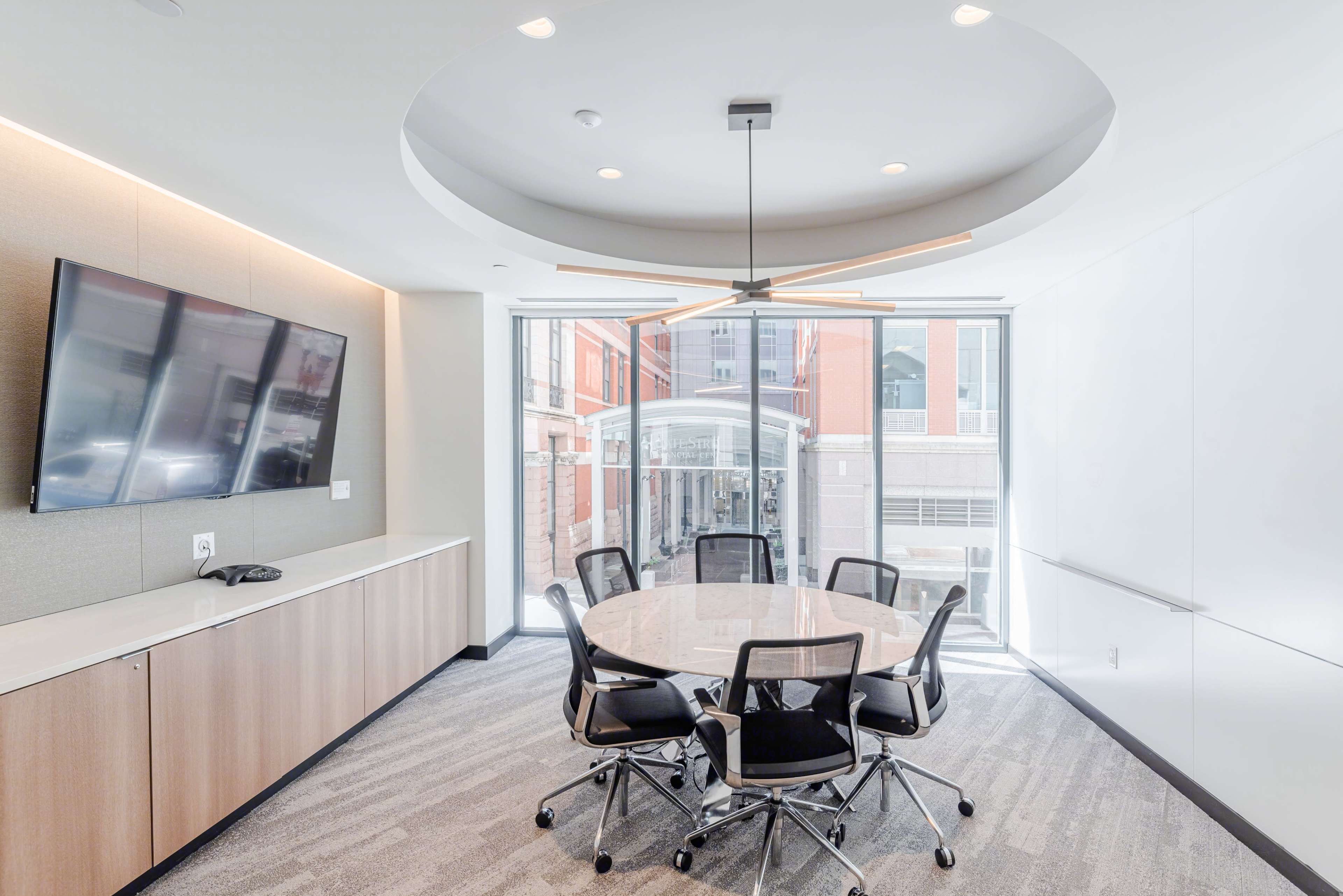 A modern conference room features a round table surrounded by six black chairs, with a large screen on the wall and windows offering a view of an outdoor space.