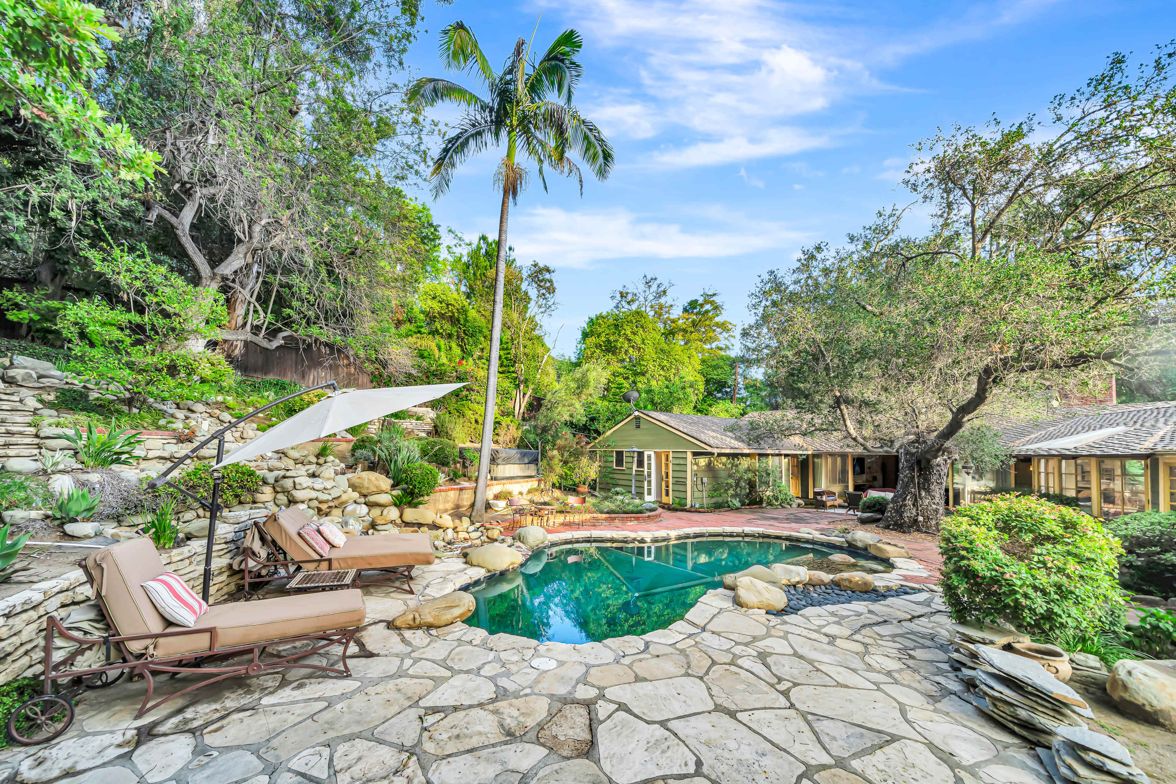 The image shows a tranquil backyard featuring a stone-paved area with two lounge chairs beside a swimming pool, surrounded by lush greenery and trees.