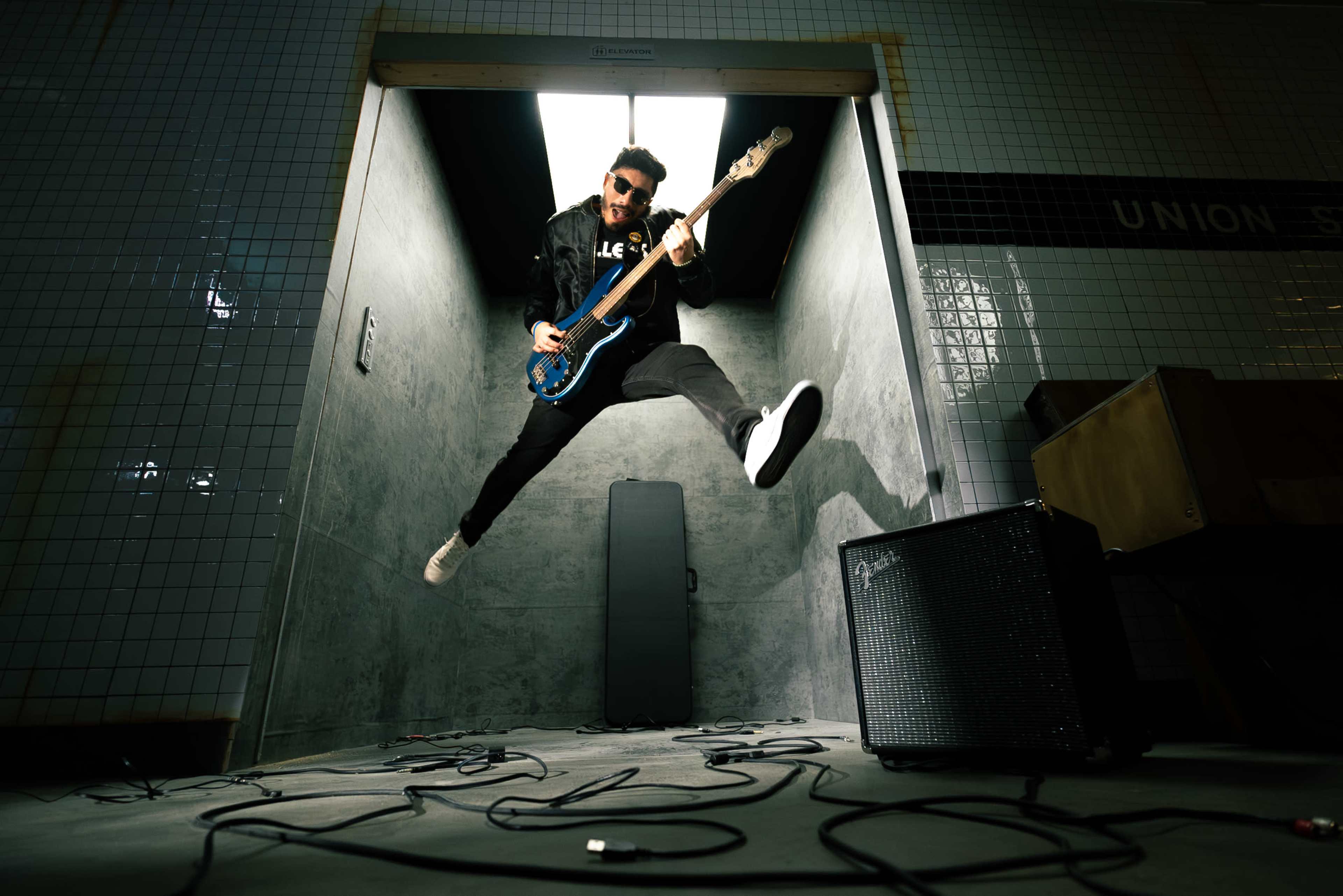 A musician jumps in mid-air while playing a blue bass guitar in a dimly lit room with tiled walls.