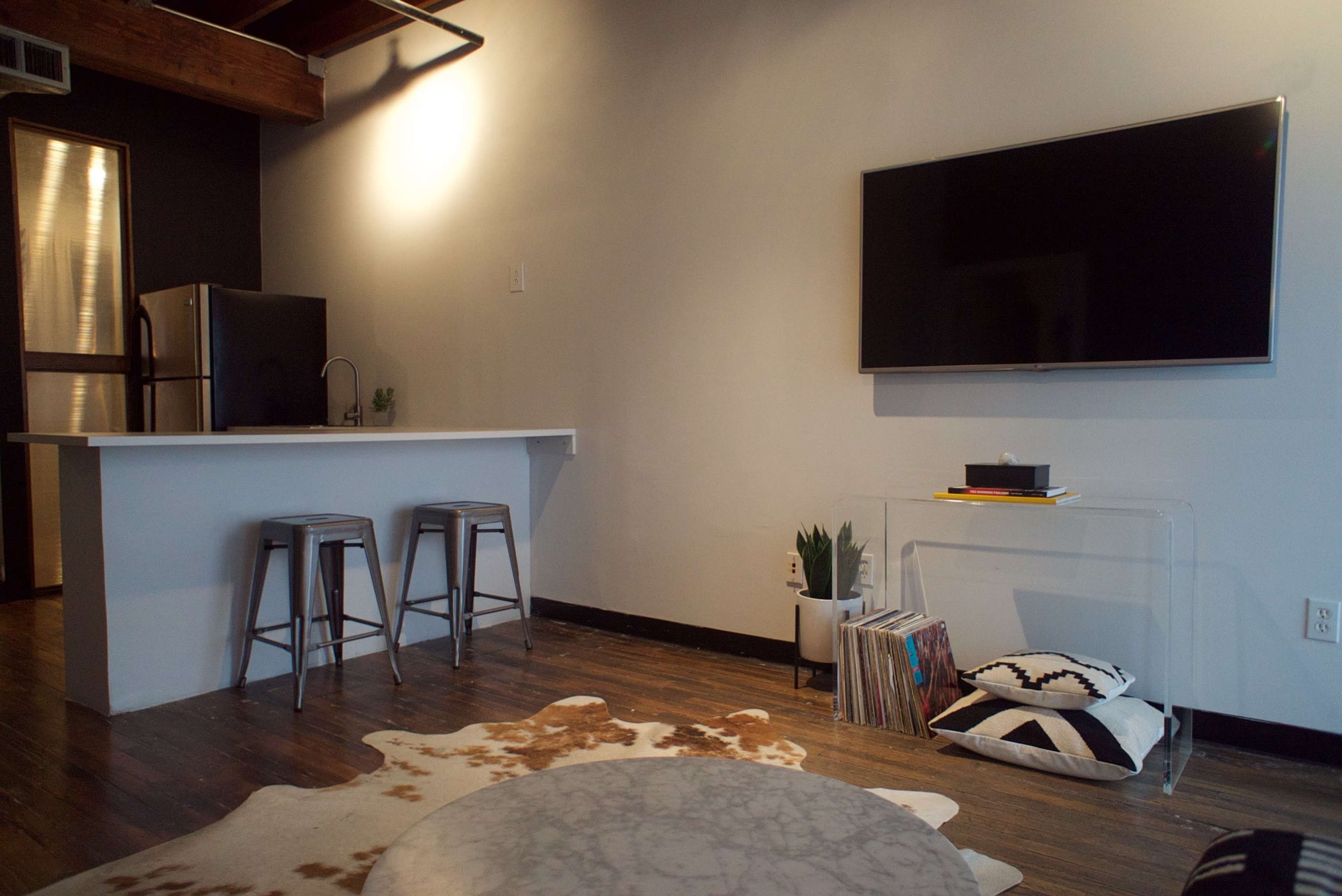 The image shows a modern living room and kitchen area featuring a minimalist design with a bar counter, stools, a television, and a clear storage unit.