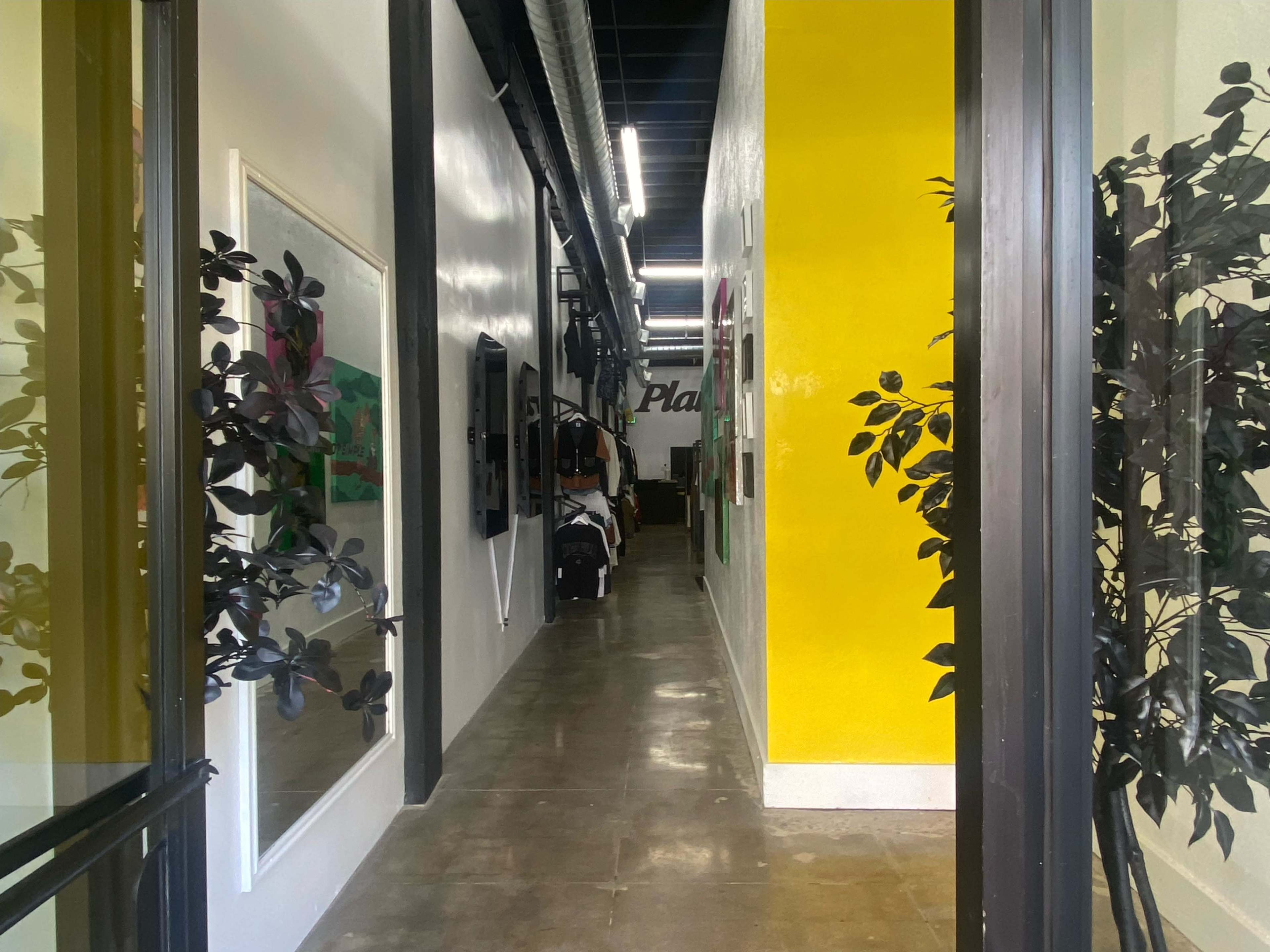 The image shows a narrow hallway lined with artwork on one side and a bright yellow wall on the other, leading to a door at the end.
