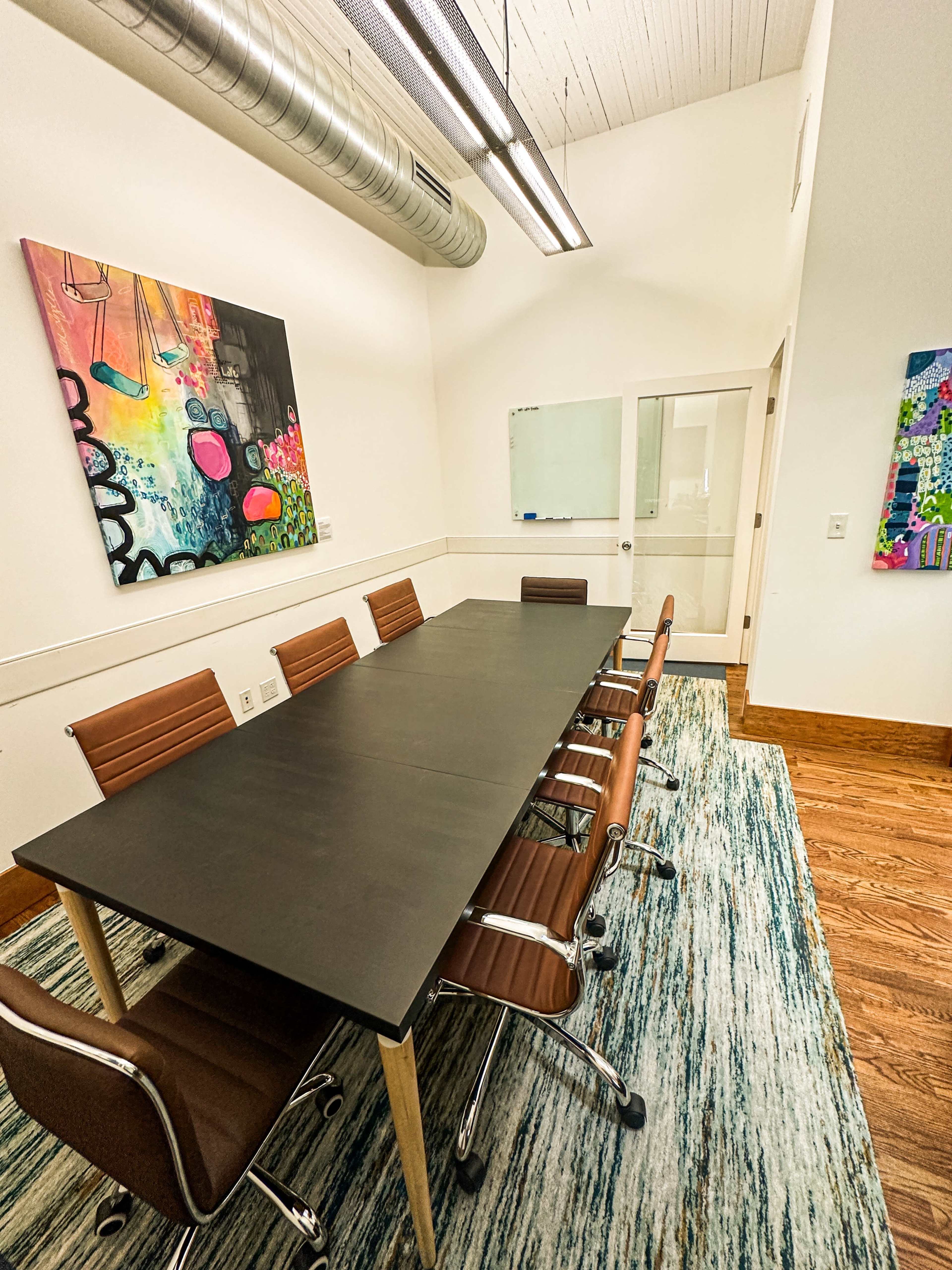 The image shows a modern conference room with a long black table, six brown leather chairs, and colorful wall art.