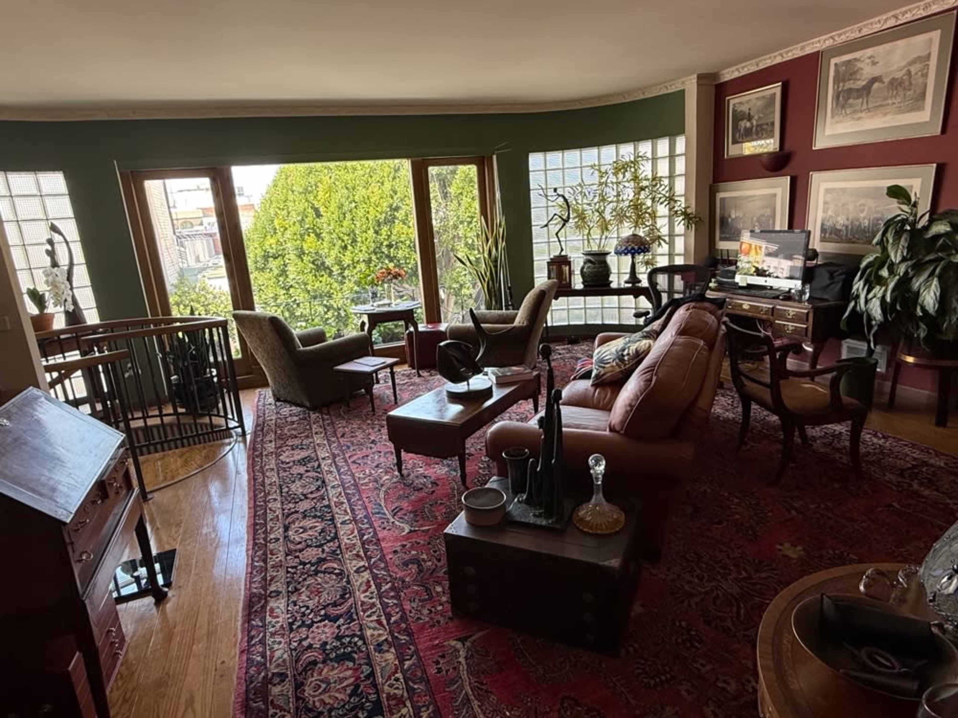 A cozy living room features a mix of vintage furniture, a large window with a view of greenery, and a patterned area rug.