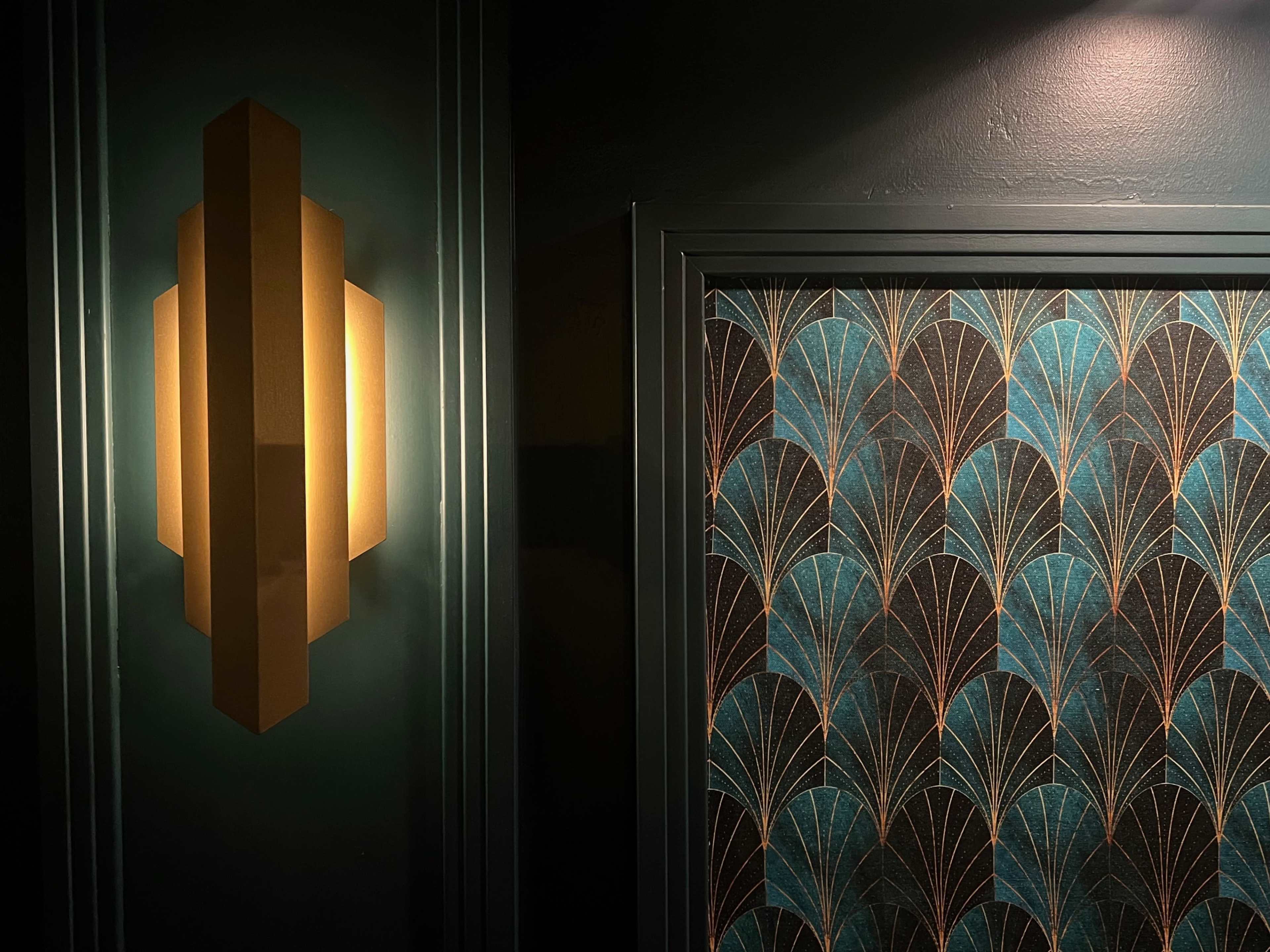 A geometric wall sconce illuminated on the left, contrasting with a patterned wallpaper of turquoise and gold fan designs on the right.