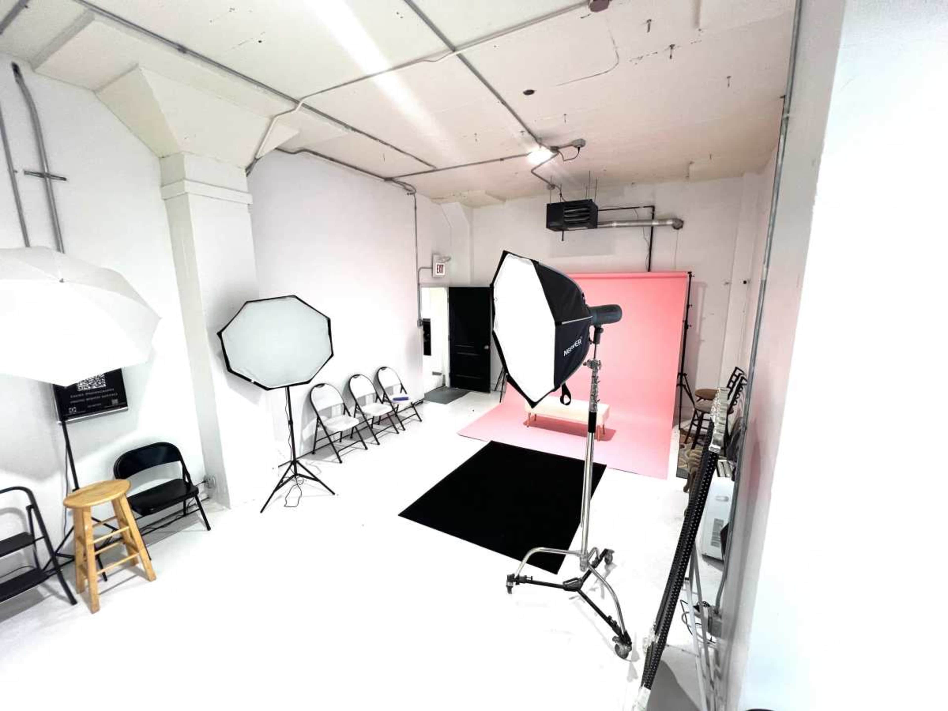 The image shows a photography studio featuring two softboxes, a large umbrella light, a pink backdrop, a black carpet, folding chairs, and a bar stool.