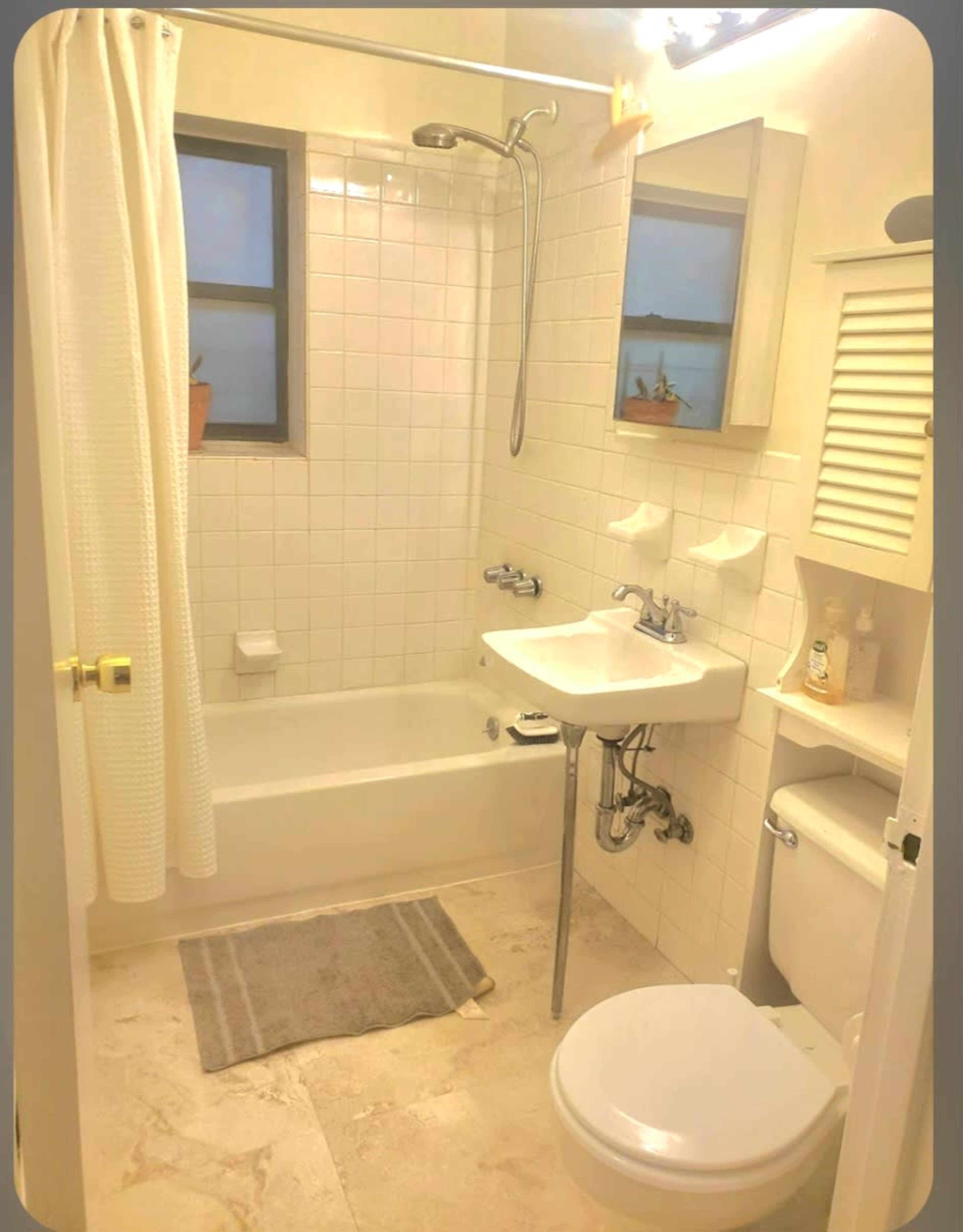 The image shows a compact bathroom featuring a bathtub with a shower above, a small sink, and a toilet, all surrounded by white tiled walls and a window.