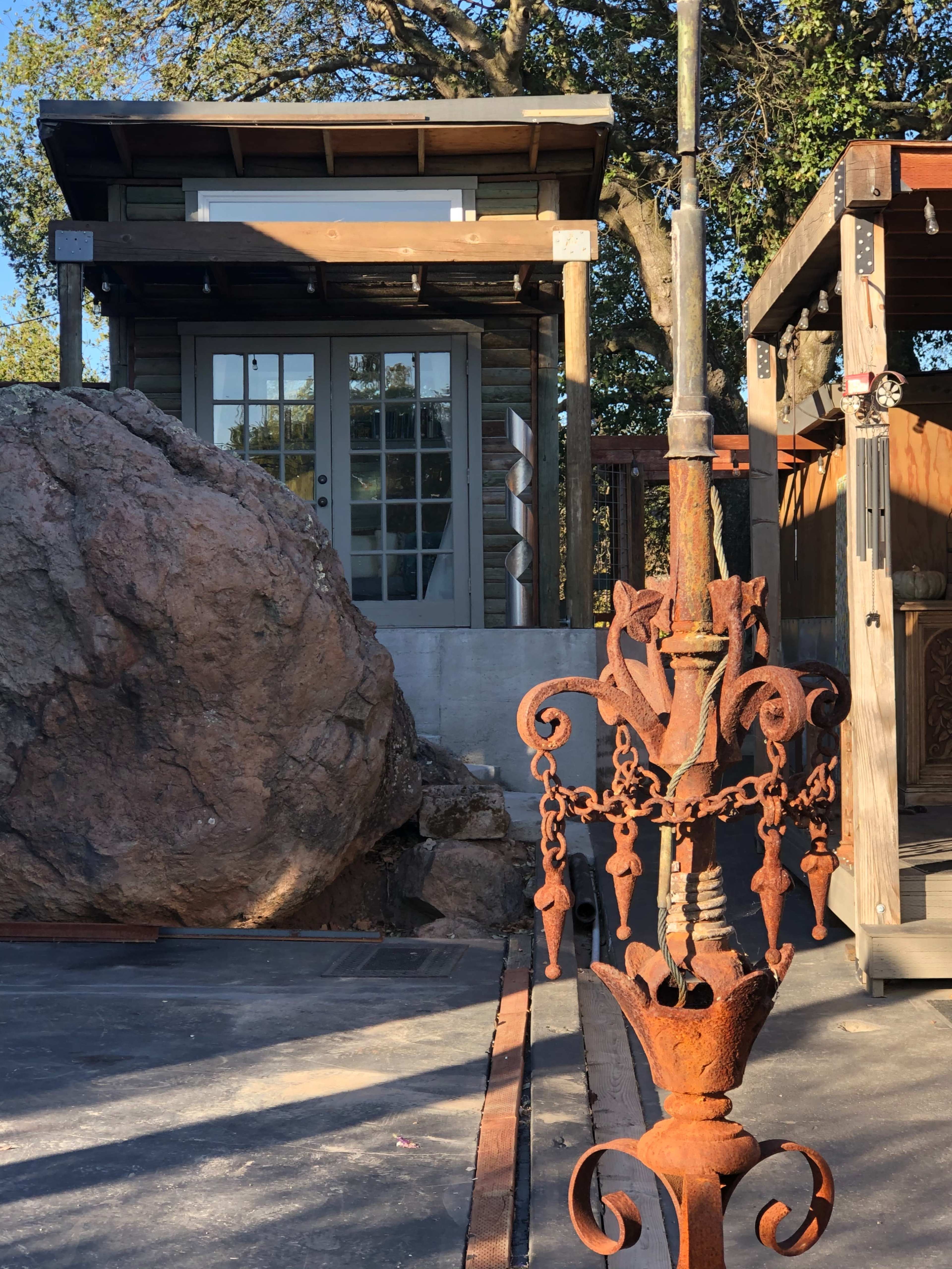 A rustic cabin with a wooden porch is situated behind a large boulder and an ornate, rusted metal post.