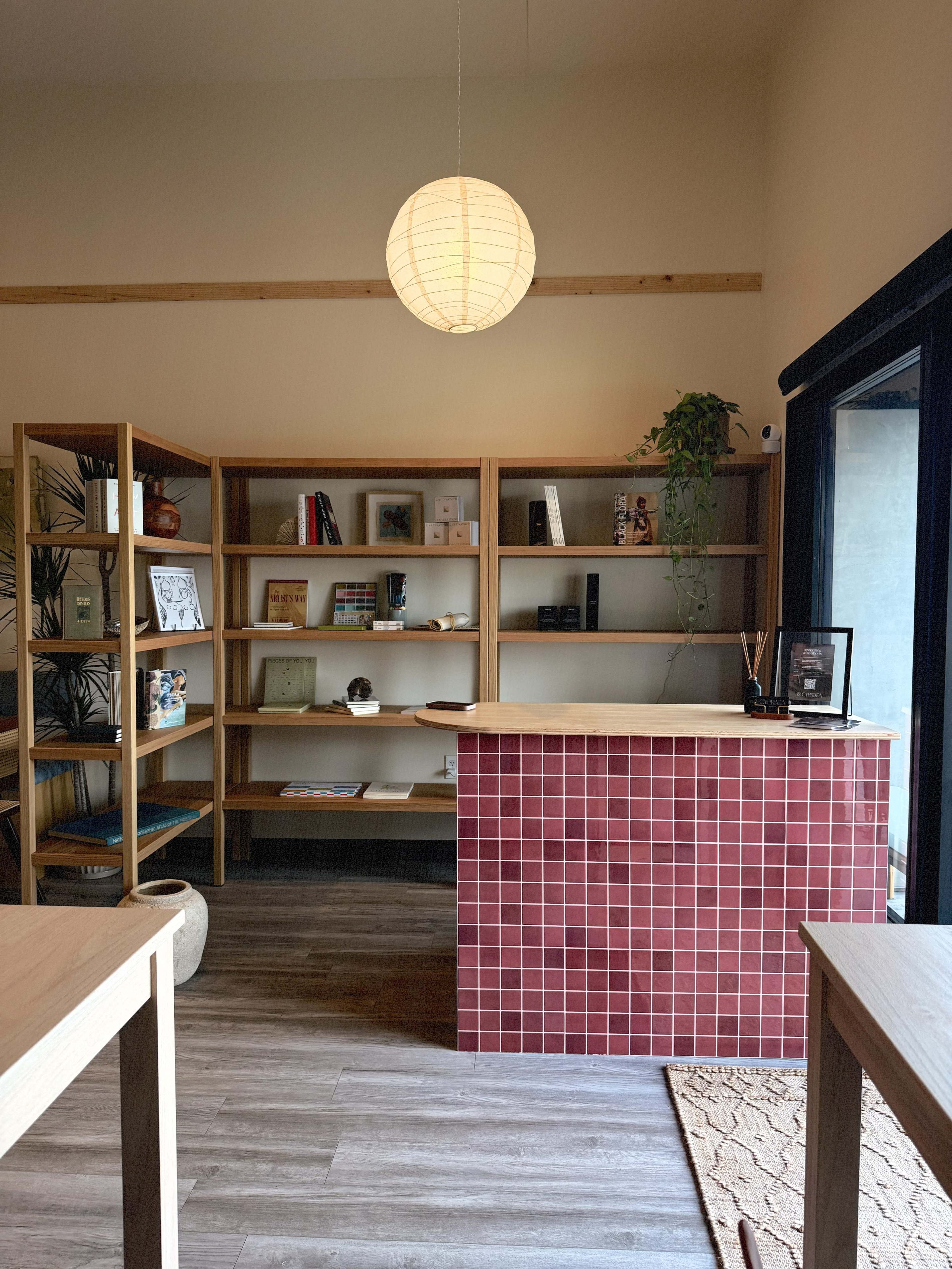 The image shows a minimalist indoor space featuring wooden shelves filled with books and decorative items, a red tiled reception desk, and a round paper lantern hanging from the ceiling.