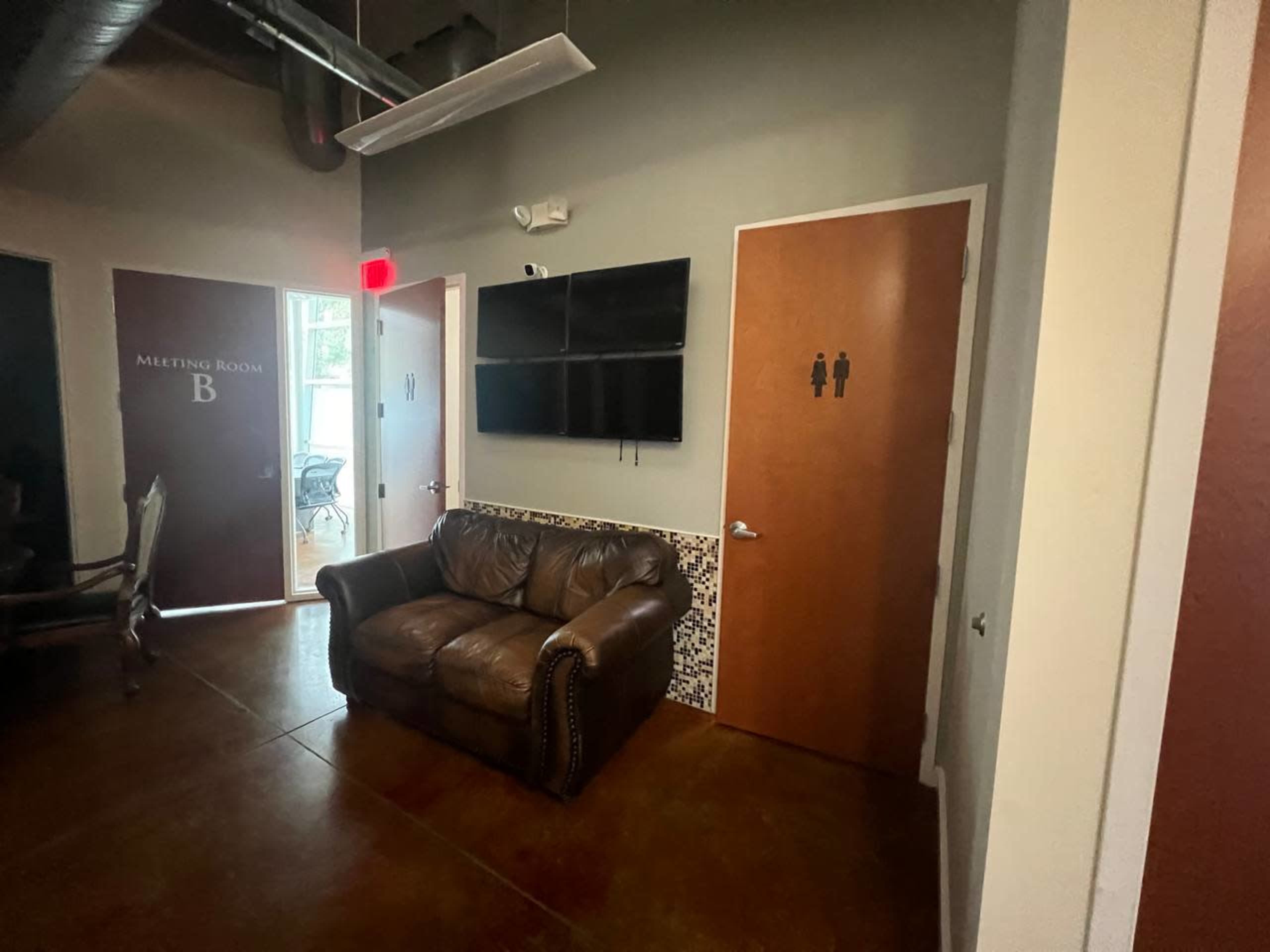 A hallway with a brown leather couch, a wall-mounted television, and doors labeled for a meeting room and restrooms.