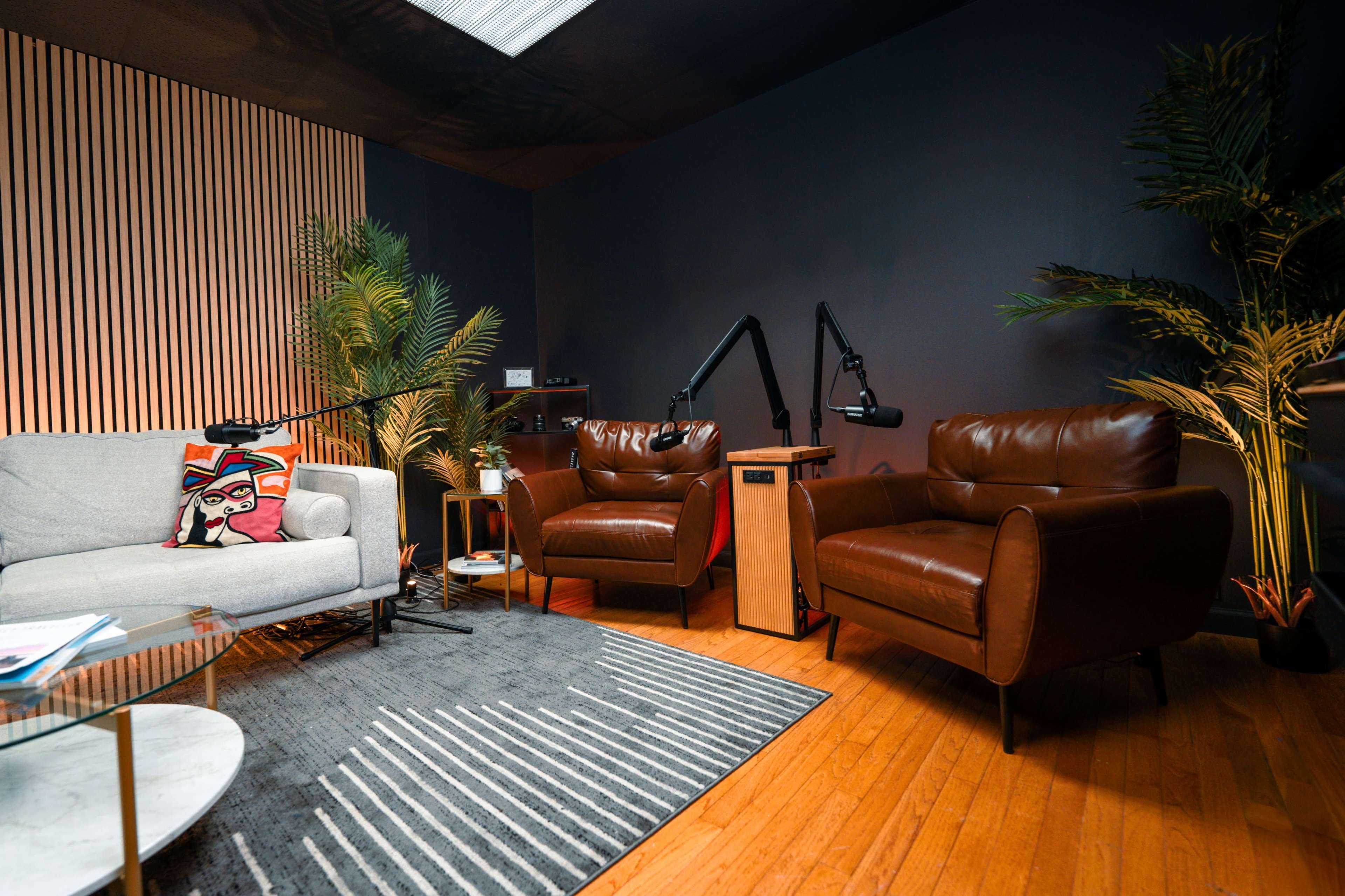 The image shows a cozy seating area with a grey sofa and two brown leather chairs on a wooden floor, accompanied by a coffee table and decorative plants.