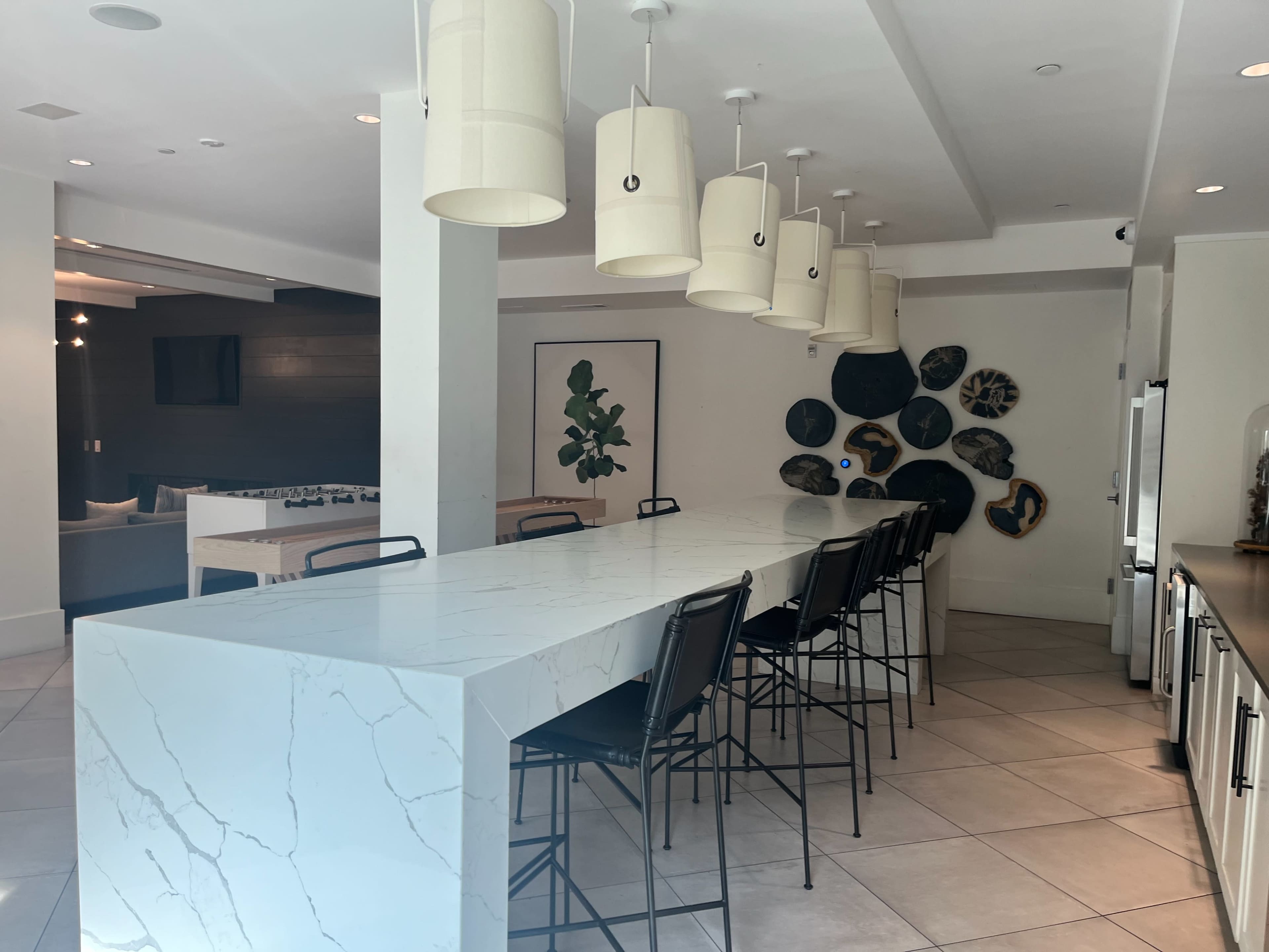 A modern kitchen area with a long white marble countertop, black bar stools, and pendant lights hanging from the ceiling.