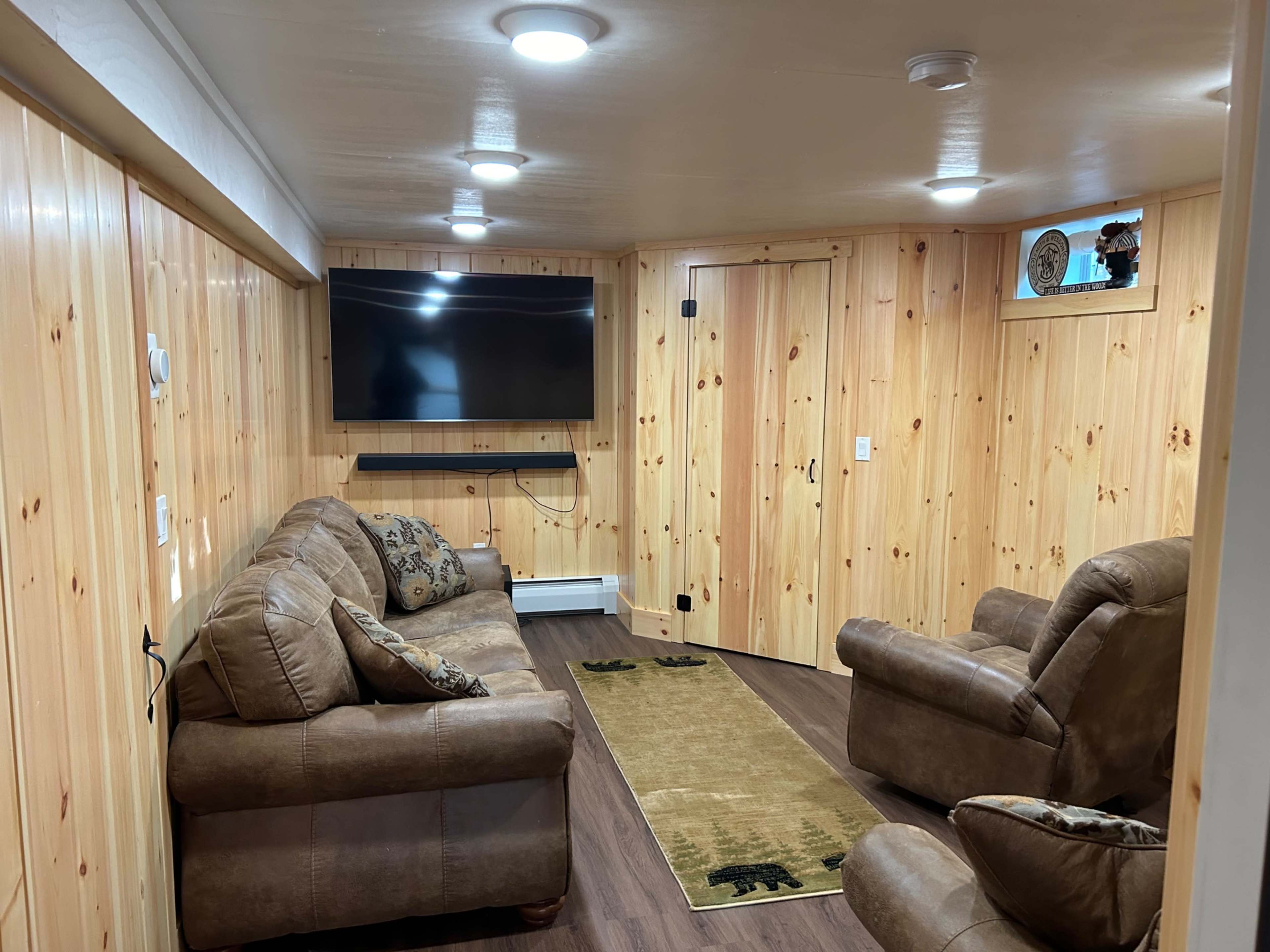 The image shows a finished basement living area with wooden paneling, a large flat-screen TV mounted on the wall, two leather armchairs, and a couch, with a rug featuring bear motifs on the floor.