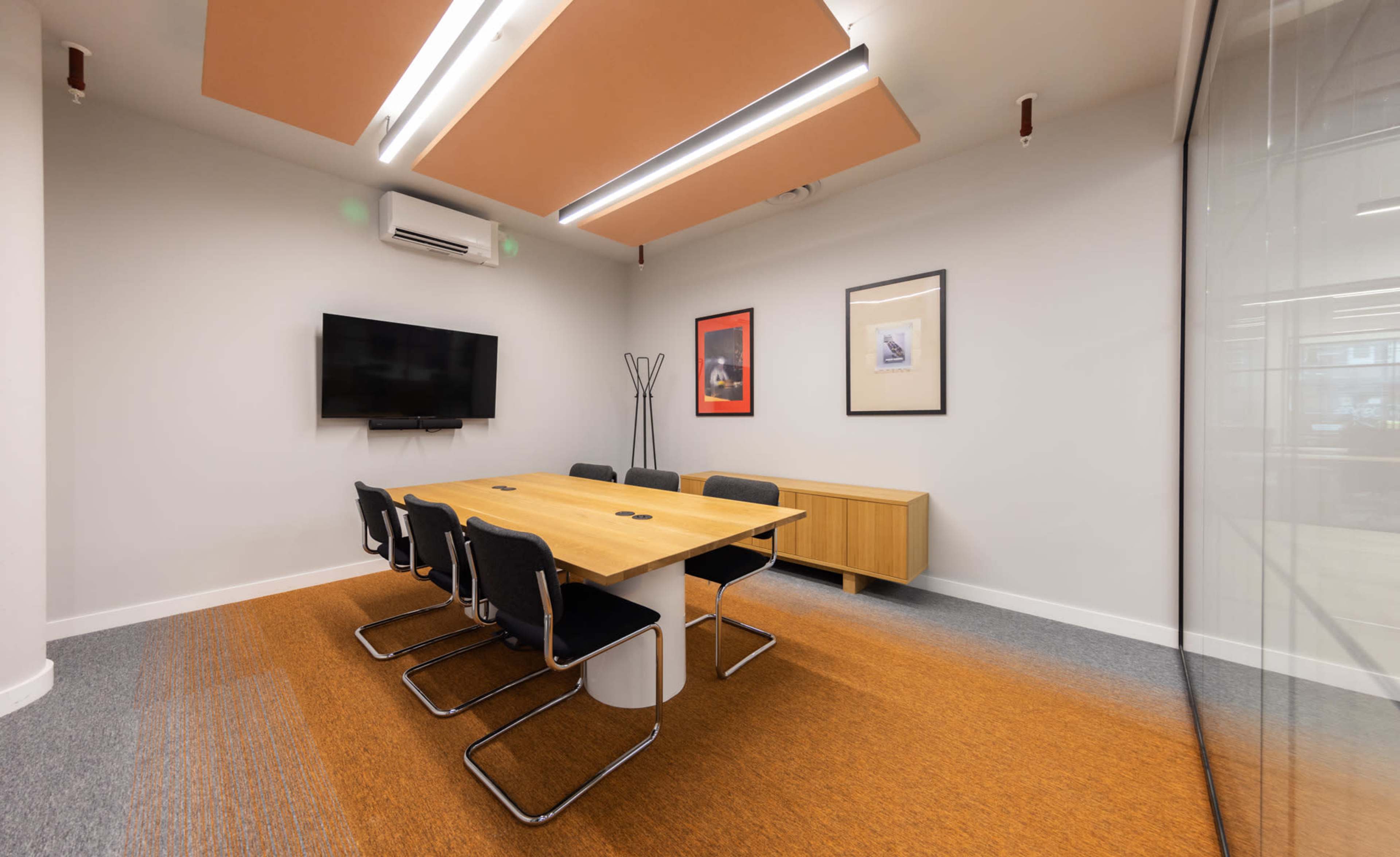 The image shows a modern conference room featuring a wooden table surrounded by black chairs, a wall-mounted TV, and framed artwork.
